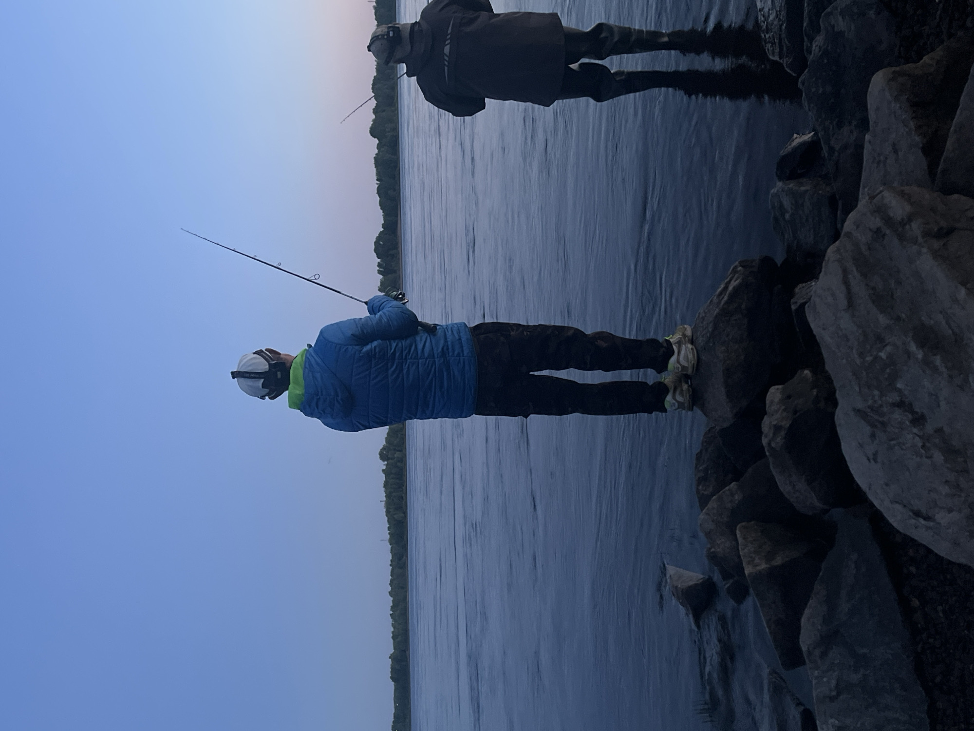 Two individuals are fishing on the banks of the Kama River in Chaikovsky, Russia, during twilight. The person in the foreground, facing away from the camera, wears a blue puffer jacket, black pants, and a white baseball cap with headphones. They are holding a fishing rod. To the right, another person, also facing away, is wearing a dark jacket and waders, and is also holding a fishing rod. They are standing on rocks near the water's edge. The river is calm, with ripples indicating a gentle current. The sky is a gradient of blue and pink, suggesting either sunrise or sunset. The background shows a dense line of trees along the opposite bank of the river. The lighting is dim, characteristic of the transitional period between day and night.