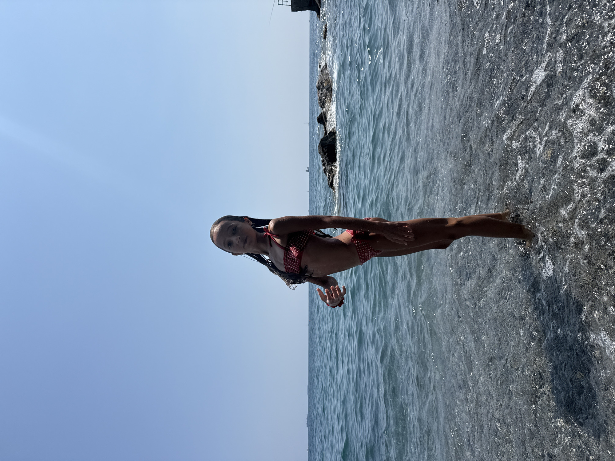 A young girl in a red and white polka dot bikini stands in the shallow water at the edge of a rocky shore. Her dark hair is wet and slicked back, framing her face as she looks towards the camera. The sun is shining brightly, casting a slight glare on the water's surface. The sky is a clear, brilliant blue, with a faint haze on the horizon. In the background, dark rocks jut out of the water, creating a rugged coastline. Further out, the sea stretches to the horizon, with gentle waves rippling across its surface. The overall impression is one of a warm, sunny day at the beach. The scene appears to be from Genoa, Italy.