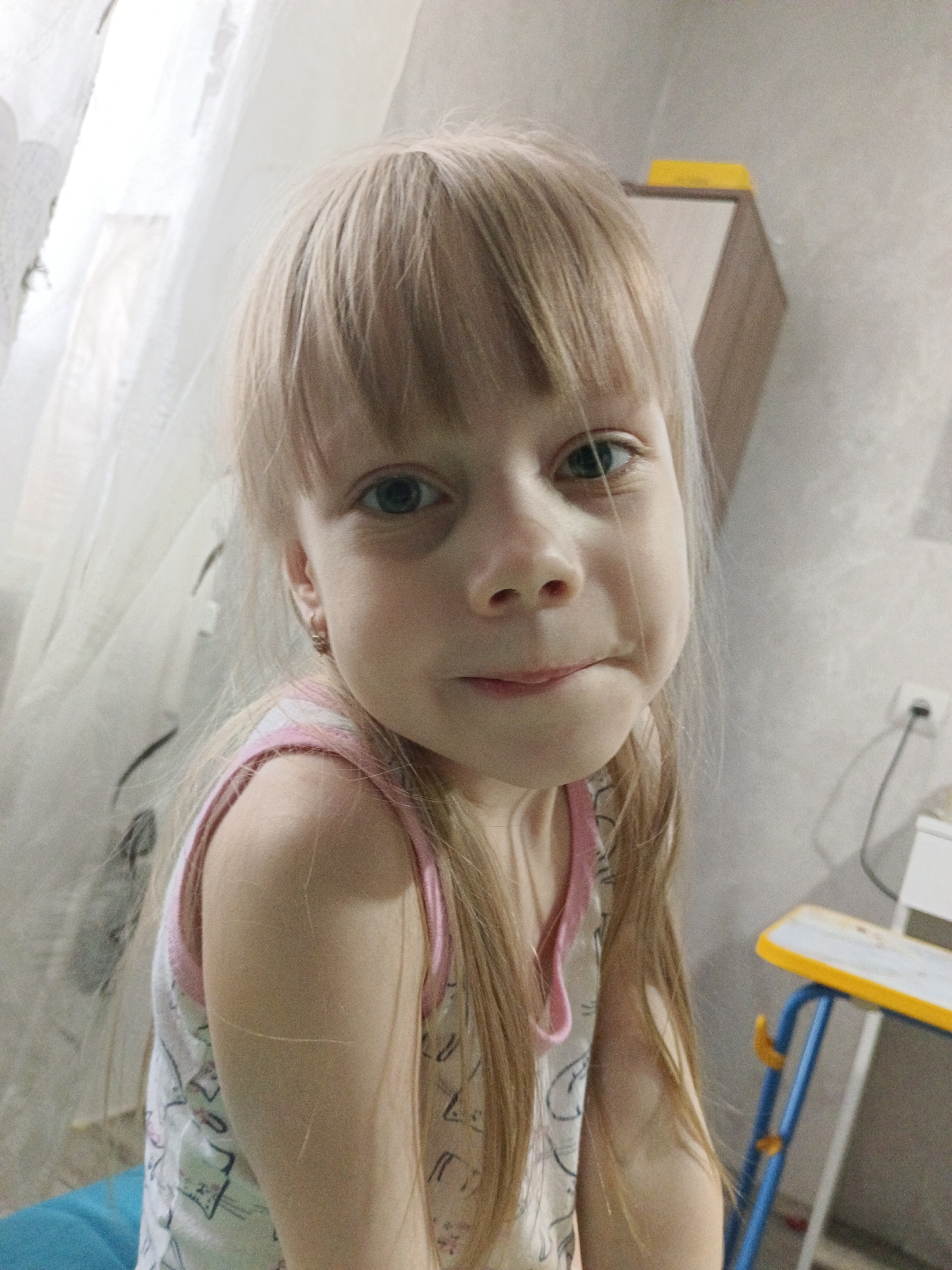 A blonde-haired girl with bangs looks directly at the camera. She is wearing a light-colored top with a pink trim. Behind her is a room with a window covered by a sheer white curtain, a wooden shelf unit with a yellow top, a light-colored wall with an electrical outlet, and a child's blue and yellow table. It is not possible to verify the location of Chaikovsky, Russia.