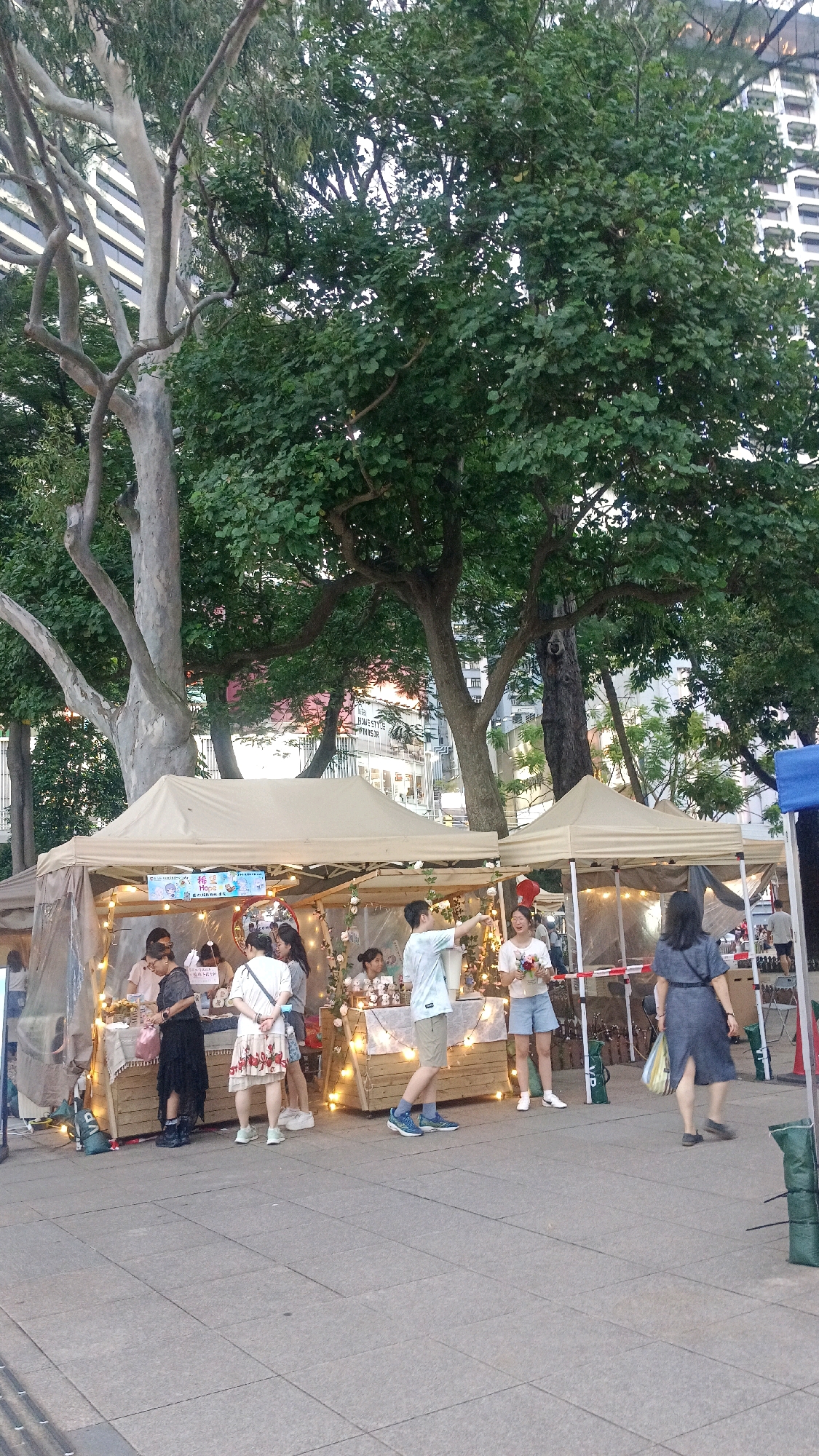The image captures an outdoor market scene, likely in Hong Kong, China, set against a backdrop of lush trees and modern buildings. Several stalls are visible, covered by beige canopies. In the foreground, a paved area is occupied by a few people.
On the left, a woman in a black dress and dark shoes stands behind a market stall, facing away from the camera. Next to her, to the right, a woman in a white and red floral dress and white sneakers also faces away, looking towards the stall. Further right, a man in a light blue tie-dye t-shirt and beige shorts, with blue and white sneakers, is seen from the side, pointing his right arm outwards with his left hand on his hip. Beside him, a woman in a white t-shirt and light blue shorts, wearing white sneakers, holds a bouquet of flowers and appears to be speaking or laughing. To her right, a woman in a dark blue dress and black shoes walks away from the camera, carrying a bag in her right hand.
The market stalls are decorated with string lights and display various items, though the specifics are not clearly discernible. One stall has a banner with Chinese characters and images. The overall atmosphere suggests a lively market day.