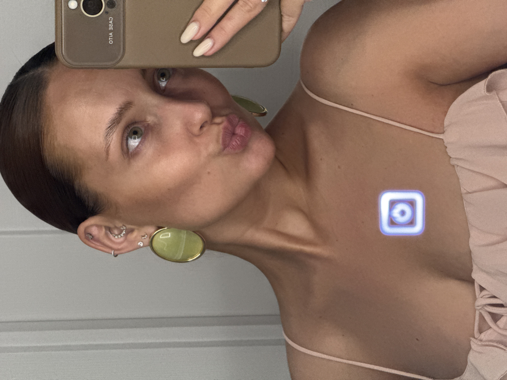 A woman is posing for a selfie. She has light skin, dark hair pulled back, and green eyes. She is making a kissing face. She is wearing a light pink top with spaghetti straps and large, round, green earrings. Her left ear has multiple piercings. She is holding a smartphone with a brown case in front of her face. The background is a plain wall. The image does not contain information that identifies the location as the city of Chaikovsky, Russia.