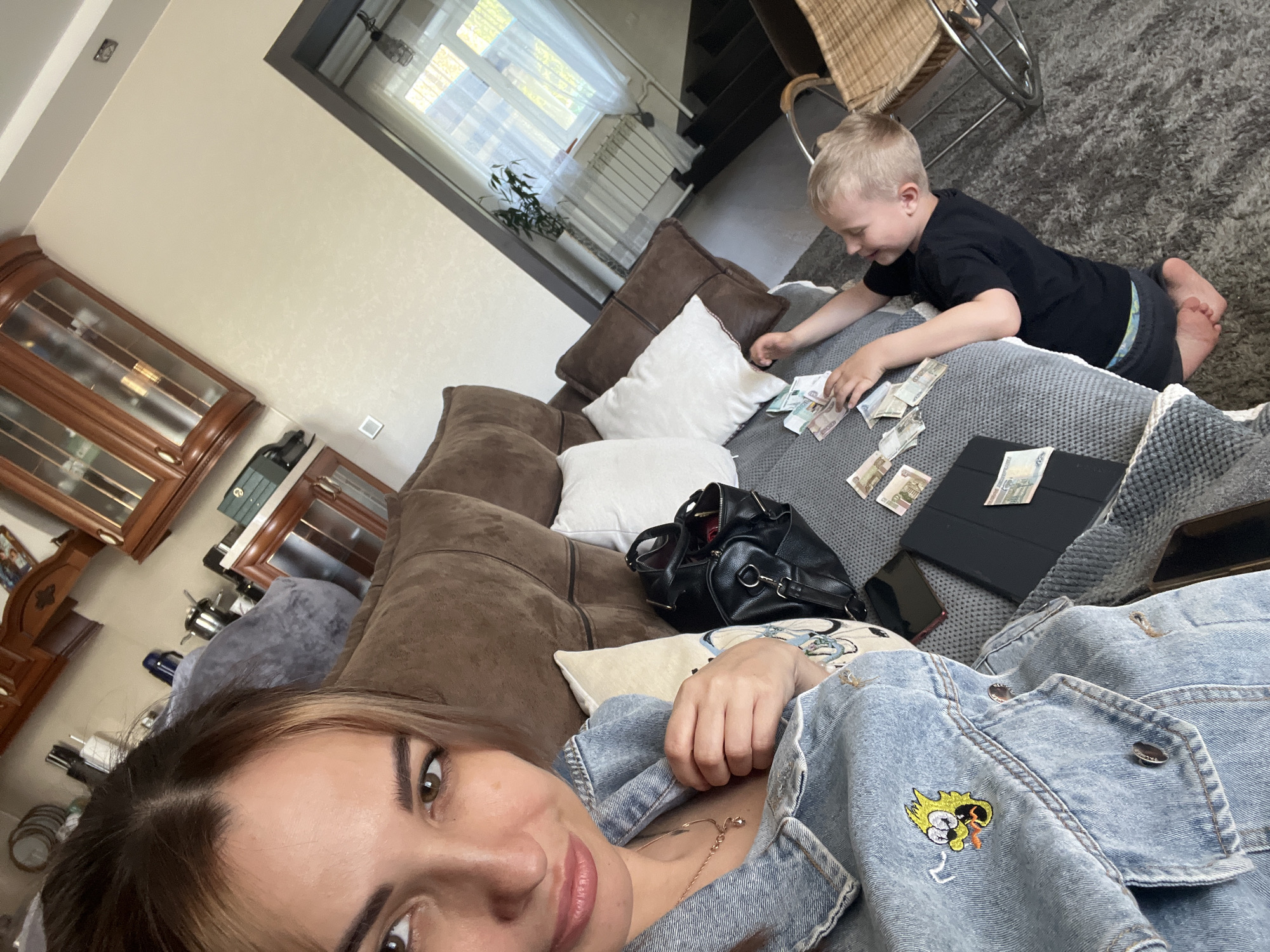 Here is a detailed description of the image:
**Scene Description:**
The image captures an interior setting, likely a living room. The shot appears to be a selfie taken by a woman who is lying on a brown couch. A young boy is present, sitting on a gray surface and interacting with numerous paper bills.
**People:**
1. **Woman:** The primary subject of the image, lying on the couch wearing a light-blue denim jacket. She has brown hair and is facing the camera, seemingly taking the picture.
2. **Boy:** A young boy with blond hair, wearing a black t-shirt. He is sitting on the gray surface next to the couch, handling paper bills.
**Objects:**
1. **Couch:** A large, brown couch dominates a section of the image.
2. **Pillows:** There are two white pillows and one beige pillow present on the couch.
3. **Paper Bills:** Numerous paper bills, possibly currency, are scattered on the gray surface.
4. **Handbag:** A black handbag sits on the couch, near the bills.
5. **Tablet:** A black tablet is seen on the gray surface alongside the paper bills.
6. **Smartphone:** A smartphone can be seen on the gray surface alongside the paper bills.
7. **Denim Jacket:** The woman wears a blue denim jacket, which features a cartoon patch.
8. **Display Cabinet:** A wooden display cabinet is located in the background.
9. **Window:** A window is visible in the background.
10. **Chair:** Woven chair can be seen in the background.
**Actions and Interactions:**
1. The woman is lying on the couch and taking a selfie.
2. The boy is sitting and appears to be counting or playing with the paper bills.
**Environment:**
The room appears to be a living space with a neutral color scheme. The furniture suggests a comfortable and lived-in environment. There is a window providing natural light, and the presence of a display cabinet hints at a domestic setting.
**Location:**
Given the interior setting and lack of specific landmarks, it's impossible to determine the city or exact location.
