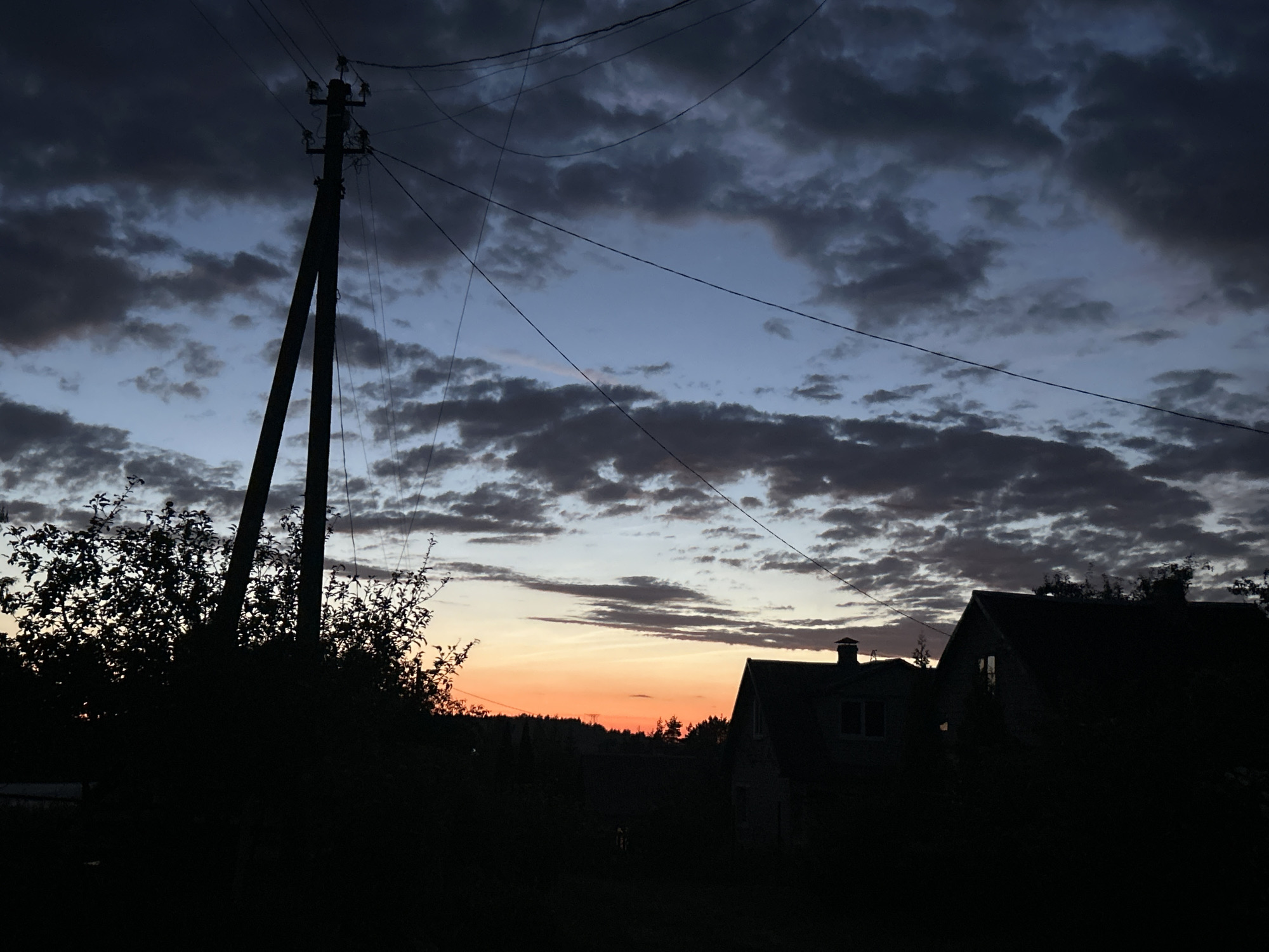 The image captures a silhouette of a rural landscape at dusk, likely within the Забалоцкі сельскі Савет, Belarus. A prominent wooden utility pole with power lines stretches diagonally across the frame, silhouetted against a dramatic sky. Dark, textured clouds fill the upper portion of the sky, transitioning to a lighter, pale blue and then a vibrant orange glow near the horizon. In the foreground, the dark silhouettes of trees and bushes obscure the ground. To the right, the dark outlines of two houses are visible, their shapes suggesting traditional rural architecture with pitched roofs. The overall scene conveys a sense of twilight settling over a quiet, possibly remote, area.