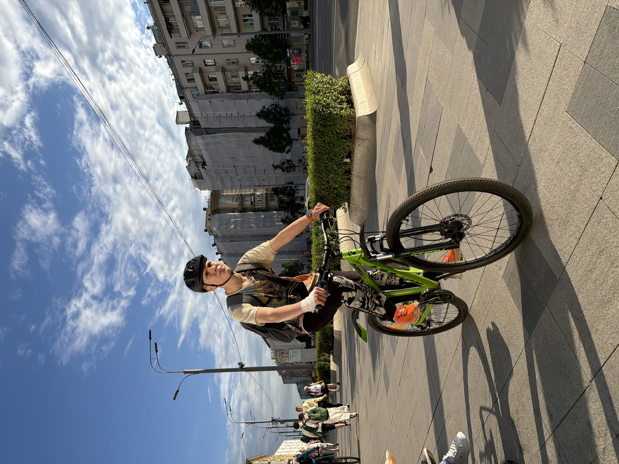 A young individual on a green bicycle is captured in an outdoor urban environment. The person is wearing a helmet, a patterned shirt, dark pants, and bright orange footwear. Their left wrist is bandaged. The background reveals city buildings under a partly cloudy sky, with overhead electrical wires visible. A line of pedestrians can be seen walking along the pavement. The ground is paved with large, gray tiles. A green hedge runs along the edge of the pavement, bordered by a concrete structure.
