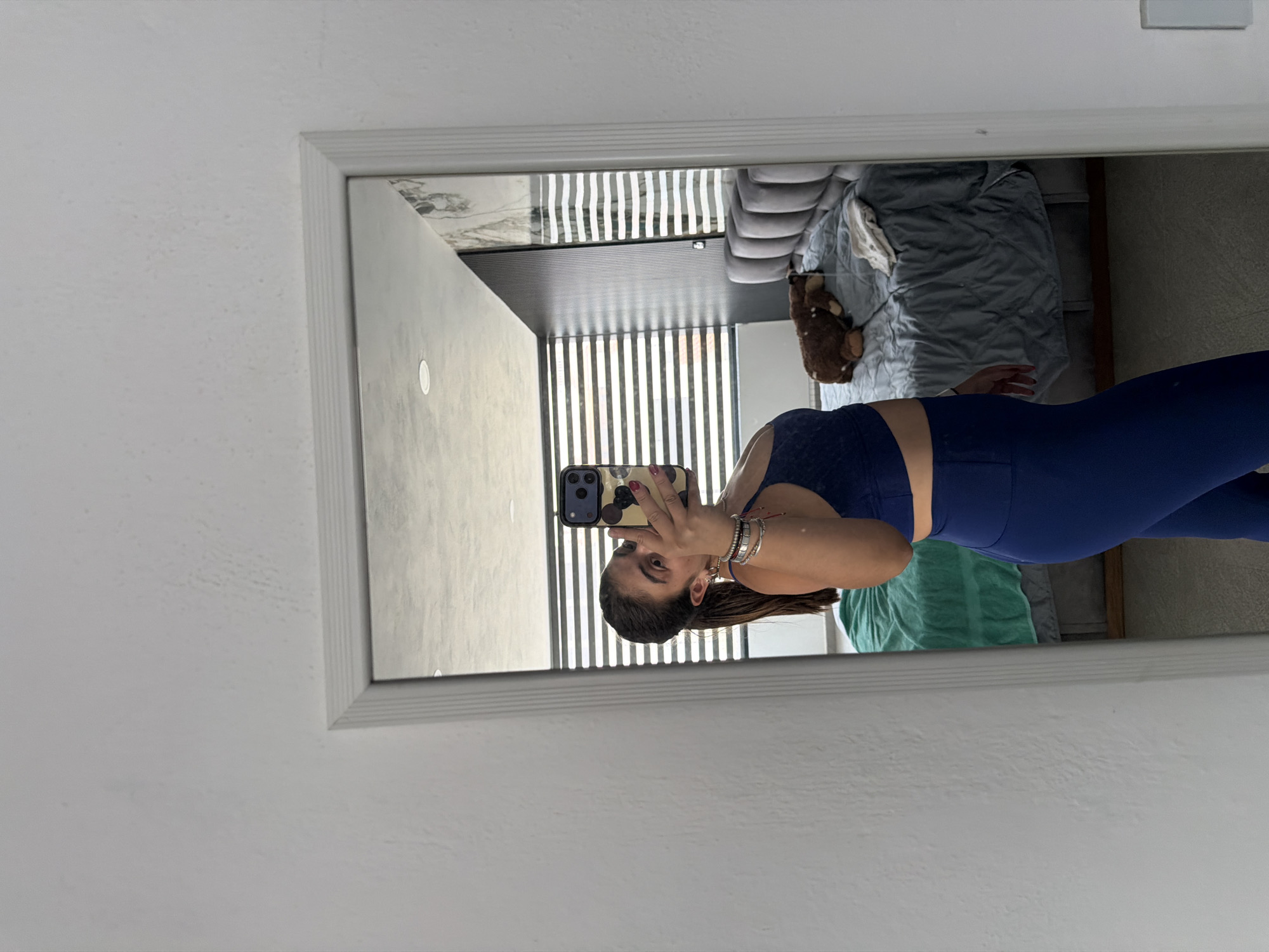 A woman in blue workout attire is taking a mirror selfie indoors. The setting appears to be a bedroom with a bed, blinds, and a light-colored wall. The location is Puebla, Mexico, but this is not discernible from the image itself, however, this information was given.