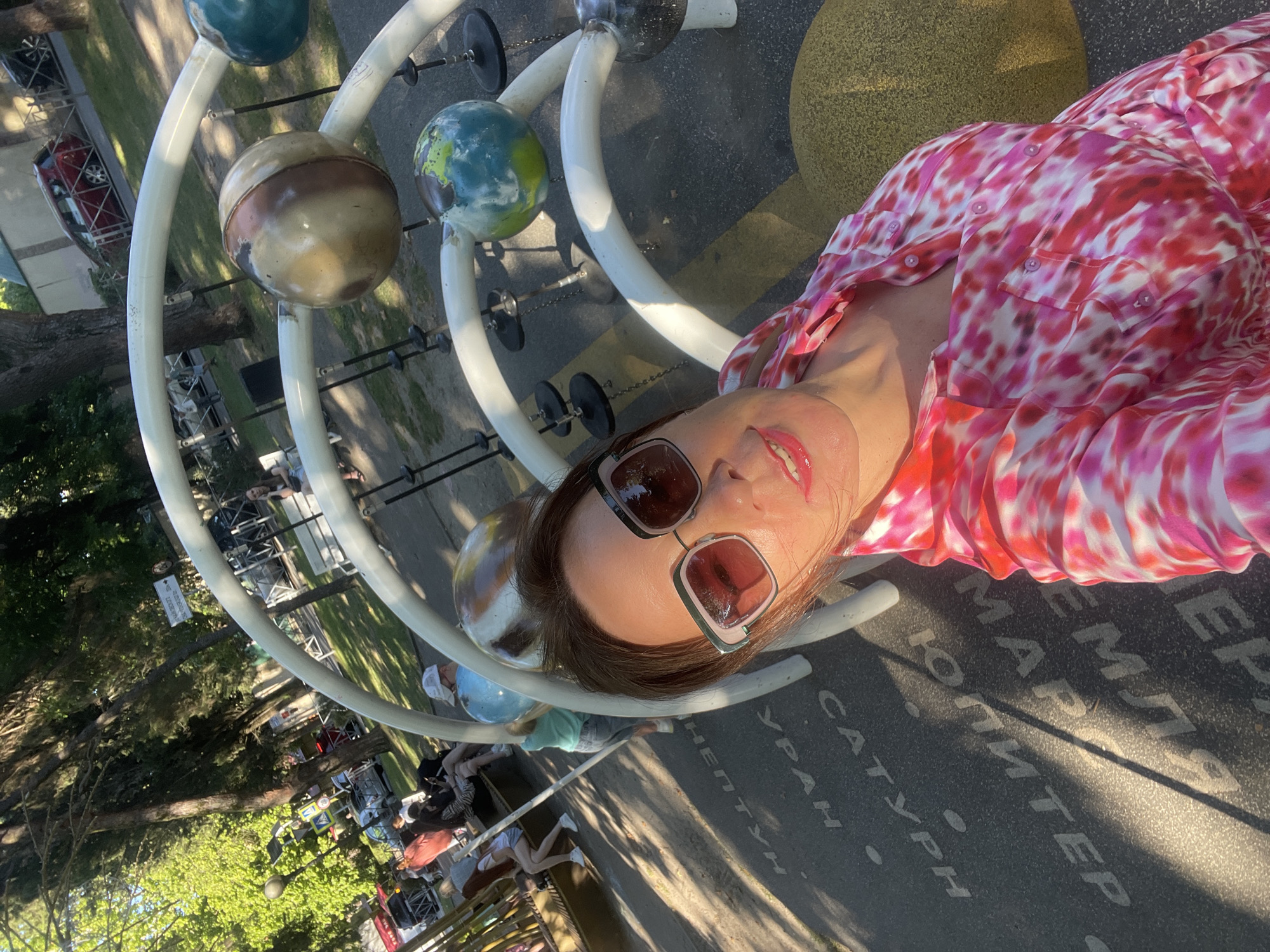 In an outdoor public space in Sochi, Russia, a woman takes a selfie. She is wearing large, square, pink-tinted sunglasses and a pink and white tie-dye print shirt. The ground beneath her features an educational display about the solar system, with text in Cyrillic indicating planets. Sunlight casts shadows from trees and a solar system model onto the ground. In the background, several people are visible, some sitting on a bench and others walking. There is also a playground structure and trees lining the area, with cars parked in the distance.
