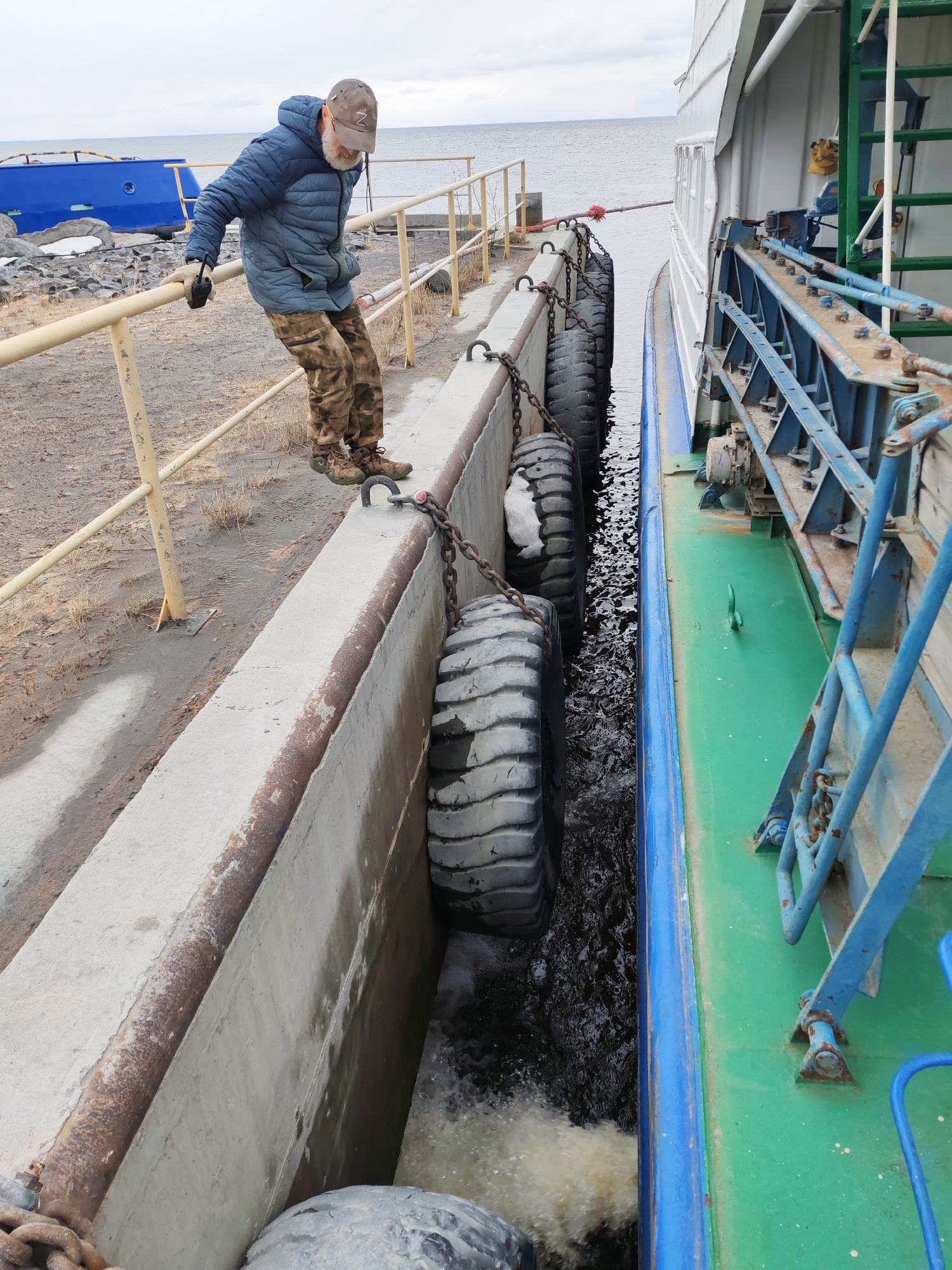 An outdoor scene depicts a man standing on a concrete dock next to a moored vessel under an overcast sky. The man wears a blue puffer jacket, camouflage trousers, brown hiking boots, a beige baseball cap with a white "Z" logo, and dark gloves. He leans on a yellow metal railing, looking down into the narrow gap between the dock and the boat, holding a dark radio or device in his right hand.
The dock is a robust concrete structure with a lower concrete wall. Large, dark, repurposed tires hang vertically as fenders, secured by chains to heavy metal cleats embedded in the dock wall, protecting the boat from impact. The vessel, painted white with a blue stripe along its hull and a green deck, is moored closely to the dock. Various blue and metal structures, including a ladder-like frame, are visible on the boat's side.
The water in the channel between the boat and the dock is dark and turbulent, with frothy white water visible at the bottom, indicating a discharge from the vessel. In the background, a large body of water, likely a sea or a large lake, extends to the horizon. On the rocky shore to the left, a blue boat hull or structure is visible, partially obscured. The ground near the dock is barren dirt with sparse dry vegetation. The scene suggests a working waterfront environment, possibly during cool weather. The location is Shcheleiki, Russia.
