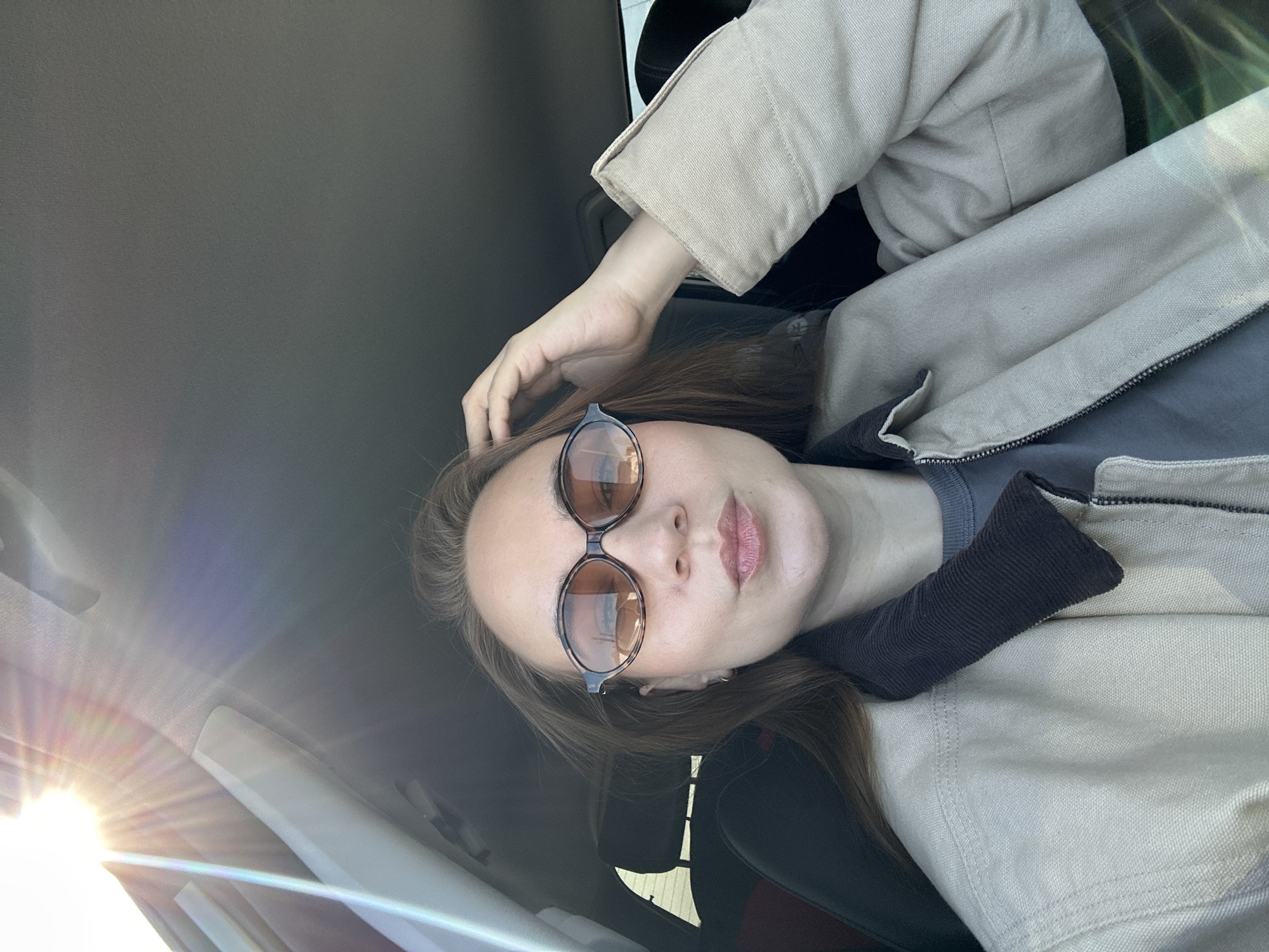 The image depicts a female individual seated inside a vehicle, likely a car, in what is identified as the city of Kharnavy, Russia. The subject, a woman with light brown hair, is positioned centrally, facing the camera. She is wearing tortoise-shell patterned oval sunglasses with amber-tinted lenses, a light beige or khaki-colored jacket featuring a dark brown ribbed corduroy collar, and a dark grey t-shirt underneath. Her left hand is raised, with fingers touching her hair on the upper left side of her head. Her lips are slightly pursed. Bright sunlight is visible in the upper left portion of the frame, entering the vehicle and creating lens flare and light streaks. Vehicle interior elements, including a light-colored headliner, an A-pillar, and a dark headrest behind the subject, are partially visible.