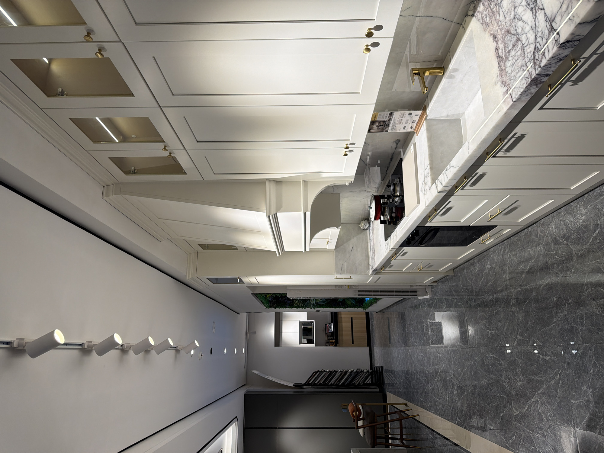 This is an interior shot of a modern kitchen, likely located in a private residence within the Chancheng District, China. The perspective is from the floor, angled upwards towards the ceiling and kitchen cabinets.
The kitchen features sleek, white cabinetry with gold hardware. Some upper cabinets have recessed lighting. Countertops are marble, possibly white marble with gray veining, extending along the wall with a gold-toned faucet and integrated sink. A stovetop is visible on the counter, with a range hood above.
Below the cabinets are drawers with horizontal gold handles. Appliances such as an oven and possibly a dishwasher are built-in. The flooring is grey marble-like tile with prominent veining.
The scene extends beyond the kitchen into other rooms. A hallway leads towards a garden view with potted plants, suggesting an outdoor area. In the foreground is a staircase with wrought iron railings and some modern art pieces. The walls are painted in a neutral white or off-white tone.
Along one wall, there is track lighting with several cylindrical spotlight fixtures. The lighting illuminates the space and highlights the modern design aesthetic. No people are visible.