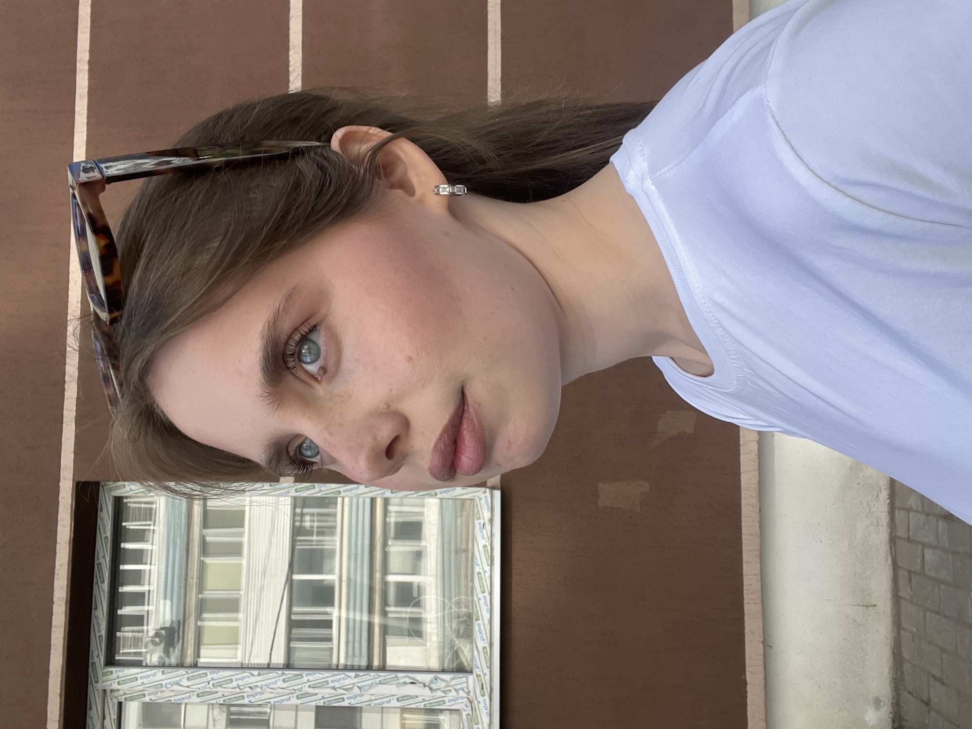 A young woman with fair skin, light brown hair, and blue eyes is facing left, with her head turned slightly towards the camera. She has sunglasses perched on top of her head. She's wearing a white t-shirt. The background shows a brown wall with white vertical stripes and a white-framed window reflecting a building. This photo was taken in Chaikovsky, Russia.