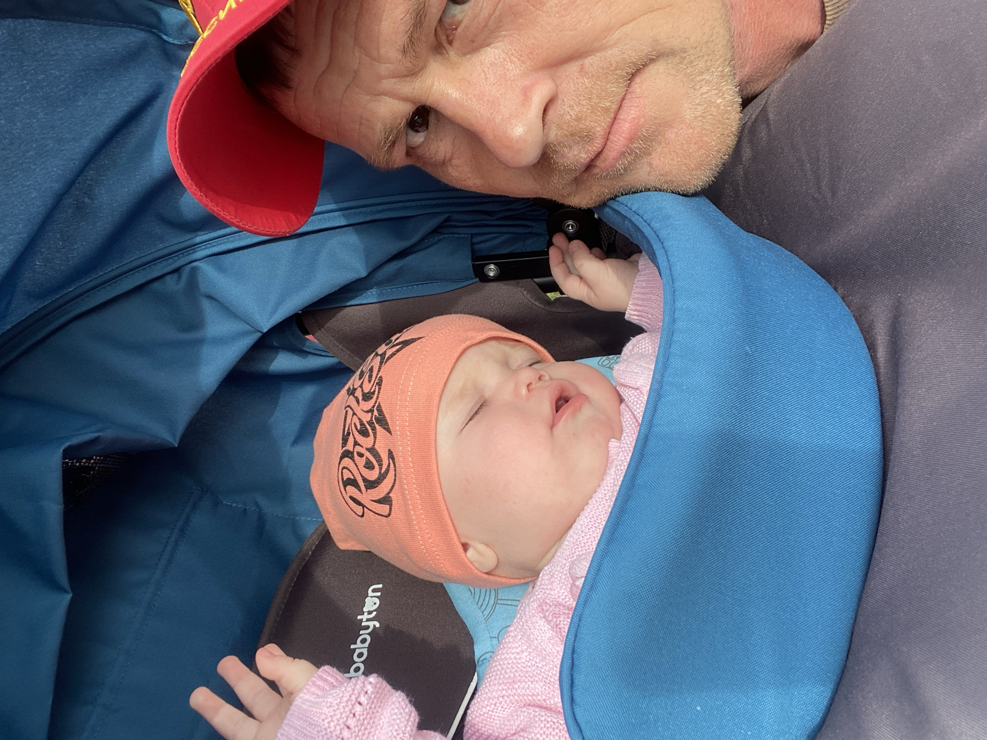 A medium shot depicts a man and a baby inside a baby stroller. The man, presumably a father or grandfather, is looking down with a pensive expression at the infant. He is wearing a red cap and has light skin, graying hair, and facial hair. The baby, dressed in pink sweater, is lying in the stroller. The baby is covered by a blue blanket. The interior of the stroller is also blue. The brand "babyton" is visible on the stroller. The location is likely outdoors, as suggested by the natural lighting.
There is no visible evidence of the image's location being in Фоки, Russia.