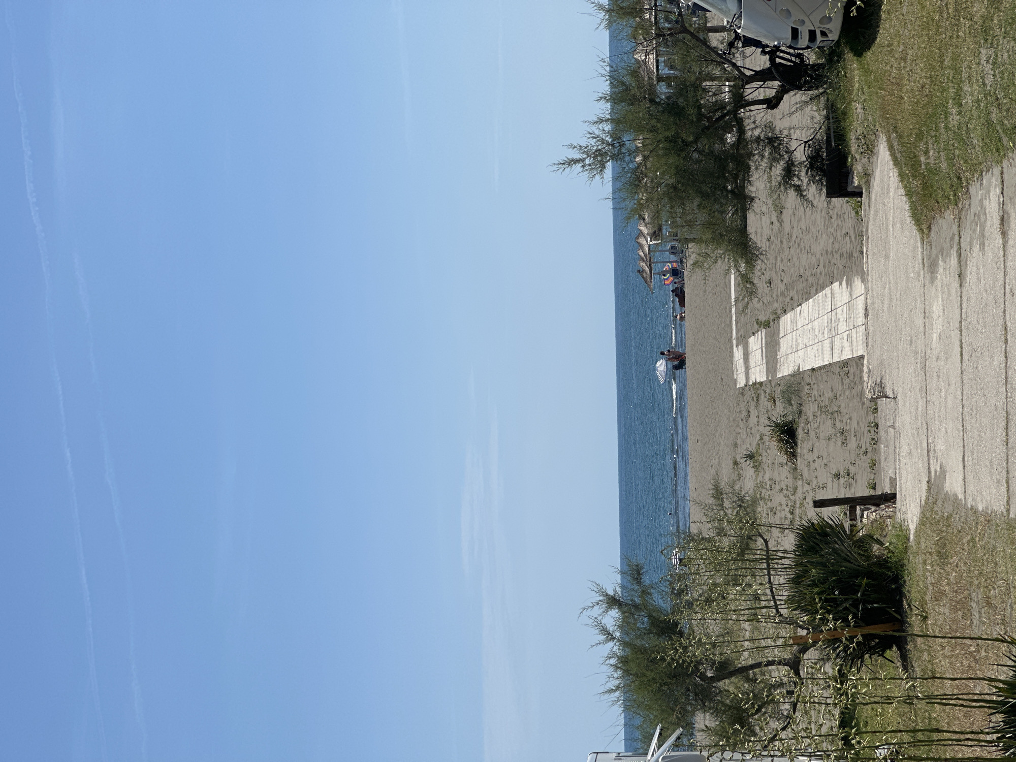 The image shows a scene at the beach in Donji Štoj, Montenegro. The sky is clear and blue. The sea is also blue, and there are people in the water and on the beach, some under umbrellas. A sandy beach separates the sea and a paved area, alongside which grass and shrubs grow. A parked white campervan is partially visible.