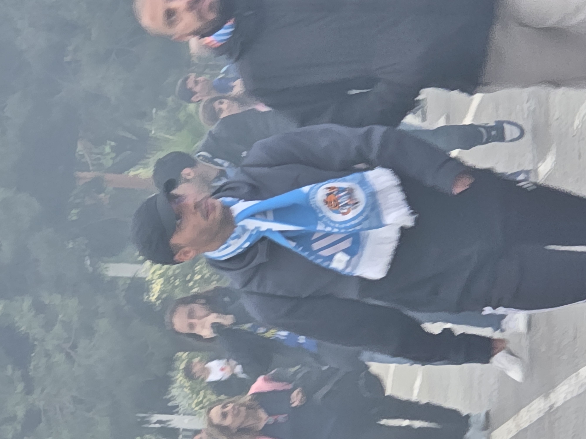 The image depicts a group of people standing on what appears to be a paved surface. A man in the foreground is wearing a dark hooded jacket, dark pants, a black cap, and a scarf with blue and white colors. Other individuals are visible in the background, some wearing scarves with similar colors. The backdrop features trees and vegetation. The location is Cornellà de Llobregat, Spain.