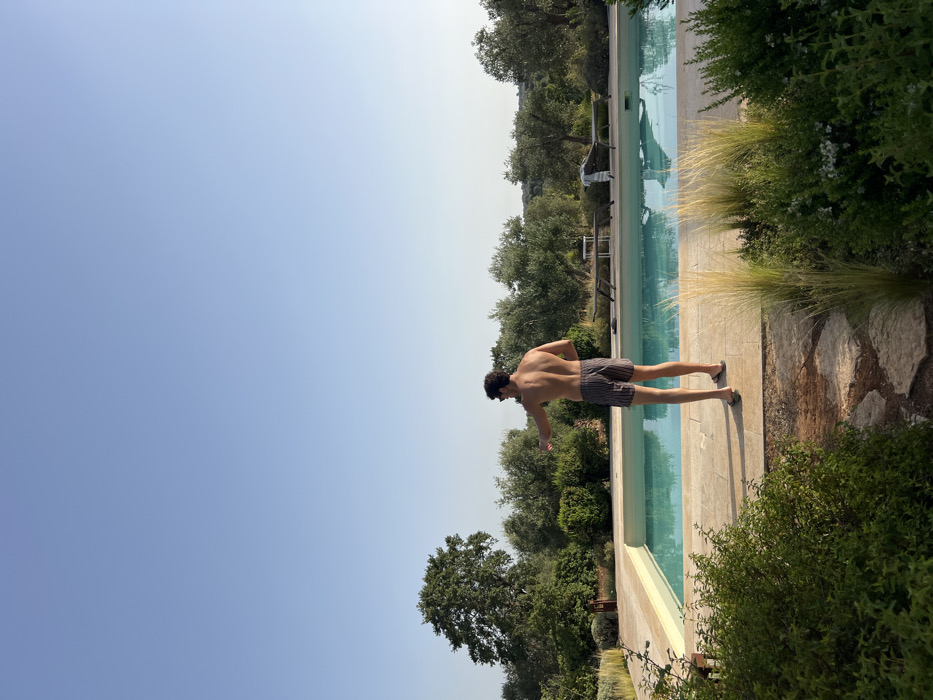 The image depicts an outdoor swimming pool area in a rural setting near Ceglie Messapica, Italy. A male subject, seen from behind, stands on light-colored paving beside a rectangular pool filled with light blue water. He is shirtless, wearing dark-striped swim shorts and flip-flops. His left arm is extended forward and slightly to the left, as if pointing. On the far side of the pool, two dark lounge chairs, one with a white striped towel, and a small white table are present on the paving. The background features an extensive landscape dominated by numerous olive trees and other green vegetation under a clear, light blue sky. In the foreground, a path of irregular stone slabs is flanked by green bushes and tall feathery grasses. The scene is illuminated by natural sunlight, casting soft shadows.