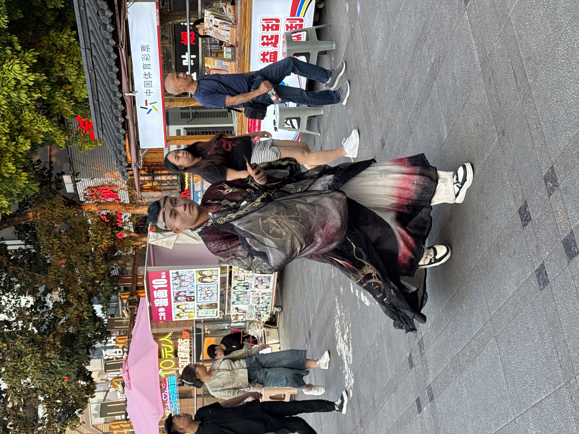 A man in a traditional Chinese robe stands on a paved street with other people. In the background are shops and signs written in Chinese. One sign advertises two-minute portraits for 10 yuan. A pink umbrella is visible on the left. The sign for "China Sports Lottery" is also visible. The location is most likely Yuzhong District, China.