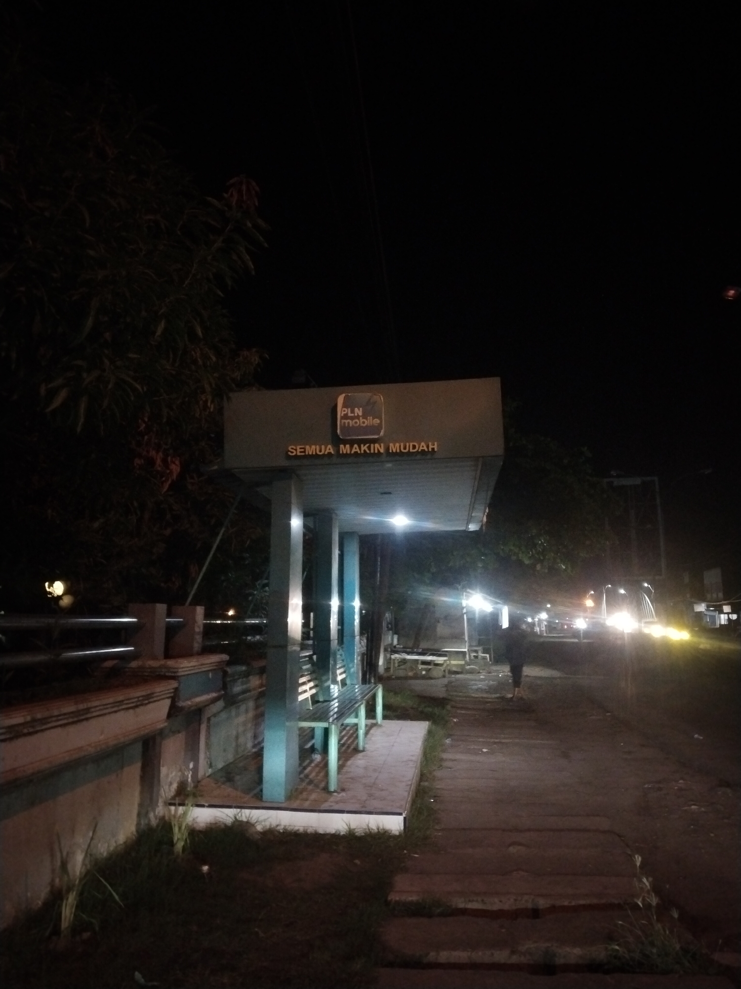 The image shows a bus stop at night in Lhokseumawe, Indonesia. The bus stop has a sign with "PLN Mobile" and "SEMUA MAKIN MUDAH" printed on it. There are benches inside the bus stop. A person is walking away from the camera on the sidewalk next to the bus stop. In the background, there are cars and street lights. A fence and some trees are to the left of the bus stop.