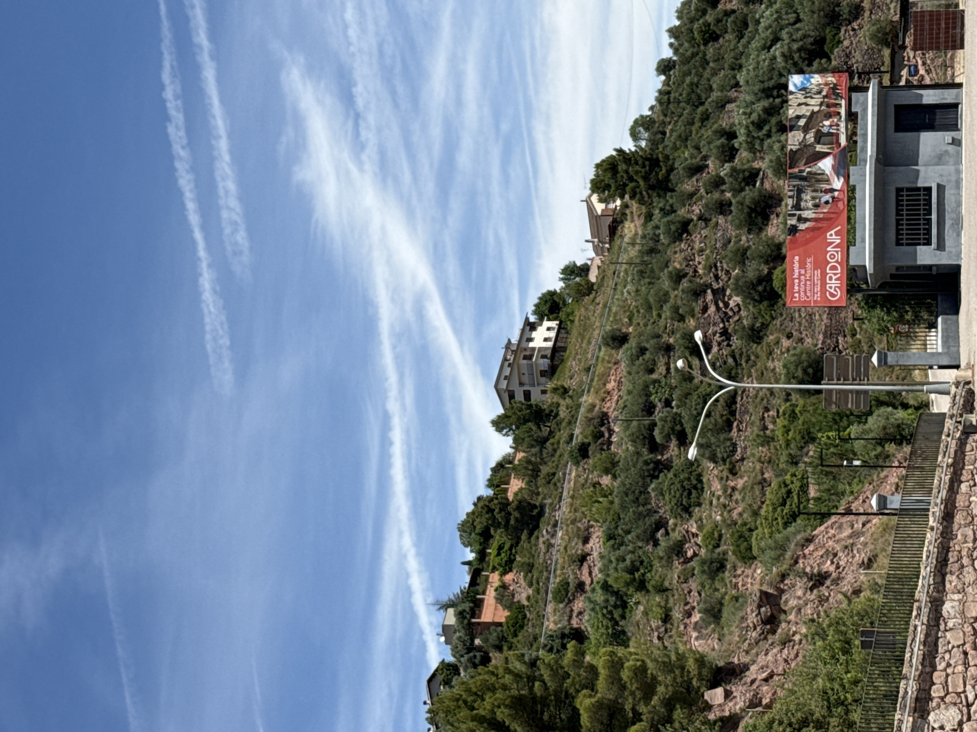 The image shows a view of a hillside in Cardona, Spain, with buildings nestled into the slope. The sky is blue with scattered white clouds. In the foreground, a stone wall and a metal fence border a road. A modern-style bus stop shelter with a red advertising banner for "CARDONA" stands in front of the hillside. The banner features images of historical architecture and people. Several streetlights are visible, including one with a distinct curved arm. The hillside itself is covered in dense green vegetation, with rocky outcrops visible in places. Several houses are scattered across the hillside.