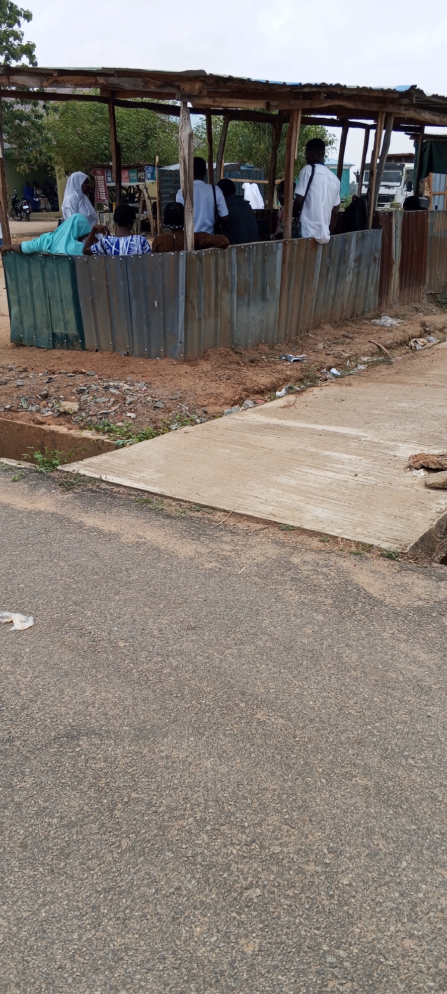 The image shows a roadside structure in Keffi, Nigeria. It appears to be a makeshift shelter or waiting area constructed with corrugated metal sheets and a wooden frame. A group of people, including at least one woman wearing a hijab, are gathered inside. Some are seated and wearing white shirts. The structure is adjacent to a paved road, and there is visible debris and dirt in the surrounding area. A concrete curb separates the dirt area from the road. The sky is overcast.