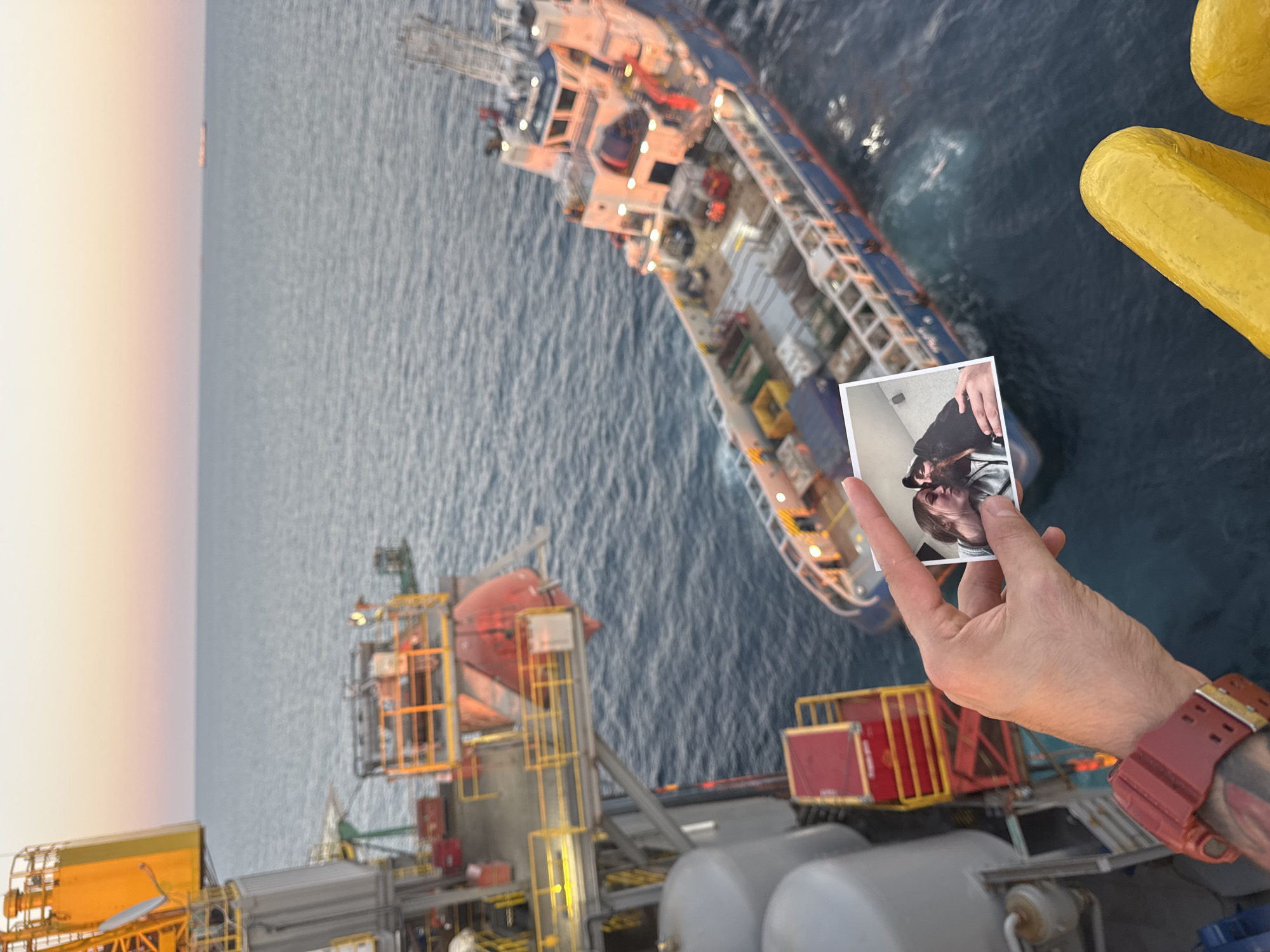 An outdoor, eye-level, medium shot captures a hand holding a photograph of a couple kissing, positioned in front of a blurred background of an offshore platform and a ship at sea during twilight. The hand, adorned with a red watch and tattoos, is reaching into the frame from the left. The photograph shows a woman with light brown hair and a man with a beard and a black baseball cap, leaning in for a kiss. Their faces are partially obscured by their heads. The background features the deep blue sea and a pale orange and pink sky. In the middle ground, a blue and red ship is visible, and to the left, a portion of an offshore platform with yellow railings and red components is present. The image lacks any discernible elements of a city, making the location of "Unknown, Italy" impossible to ascertain.