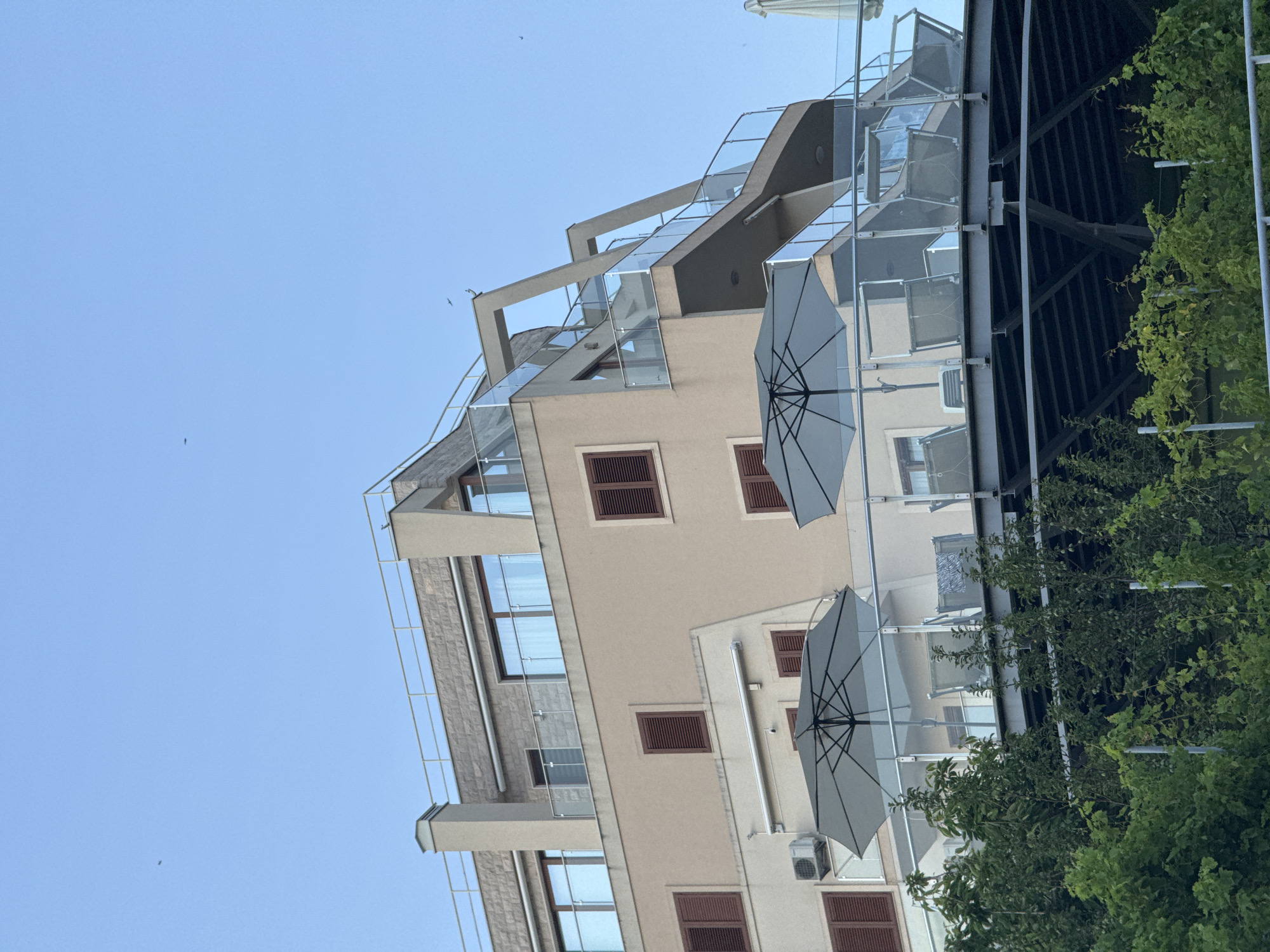 A modern, multi-story building with a beige facade and a stone-clad upper level is depicted against a clear blue sky in Boreti, Montenegro. The building features numerous windows with dark frames and wooden shutters, as well as glass balconies. Several lounge chairs and large, light-colored umbrellas are arranged on a spacious outdoor terrace in the foreground, suggesting a hotel or residential complex. Lush green foliage obscures the lower portion of the structure and the ground.