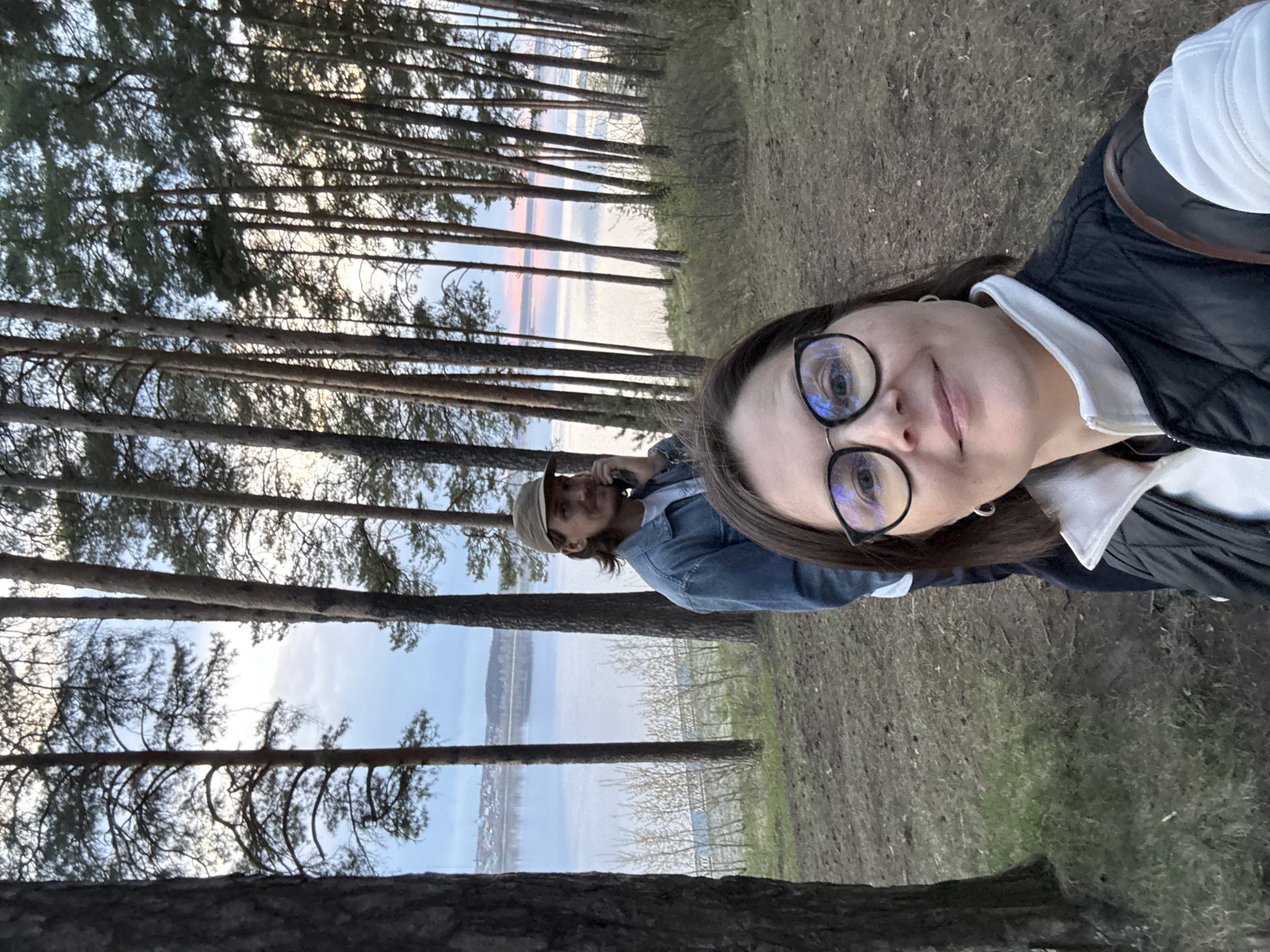 The image is an outdoor selfie taken by a person wearing glasses, a white shirt with a black vest. A second person is partially visible in the background wearing a denim jacket and tan hat. They are standing in a forest. Tall pine trees are present on both sides of the frame. Through the trees, a body of water is visible, appearing to be a river or lake. Along the opposite shore, a city is discernable. Based on the appearance of the landscape and city in the distance, this image appears to be taken in or near Izhevsk, Russia, possibly near the Izhevsk Reservoir. The ground is covered in dry vegetation.