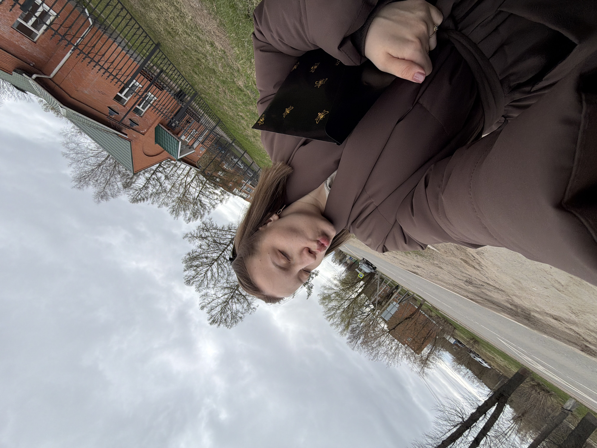 A young woman, wearing a dark brown puffer coat, is shown posing for a photograph outdoors in Reshetnikovo, Russia. She holds a black flat object, possibly a small bag or notebook, adorned with multiple golden bee illustrations. Her eyes are mostly closed, and her lips are slightly pursed in a playful expression. A ring is visible on her left hand.
The setting includes a two-story brick building with a gabled green roof and white-framed windows, partially visible behind a black ornate metal fence. Adjacent to the fence is a grassy area, and beyond that, a paved road with white markings. Bare trees, indicating a late winter or early spring season, are scattered across the landscape. The sky is overcast and grey, suggesting a cloudy daytime. No visible text is discernible on any objects or in the environment.