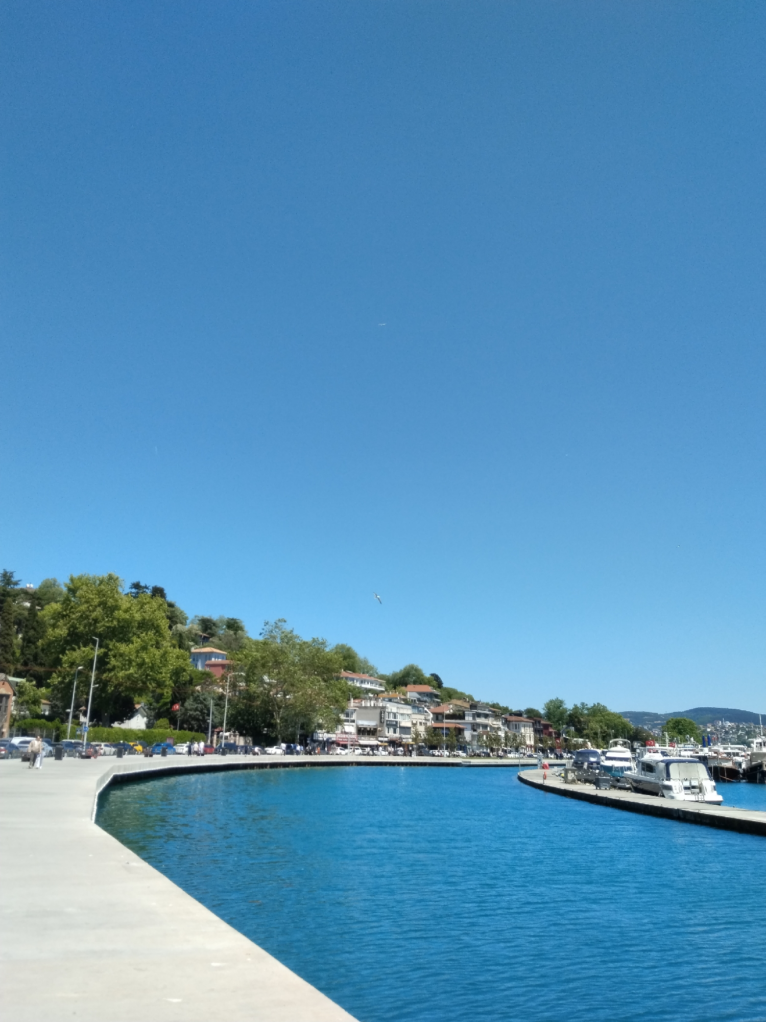The image shows a waterfront scene on a sunny day in Unknown, Unknown. A curved concrete pier extends into blue water. Along the pier, on the left, is a road where cars are parked, and a few people are walking. The opposite shore, lined with buildings and trees on a hill, forms a bay. Boats are moored at the end of a pier. The sky is clear and blue.