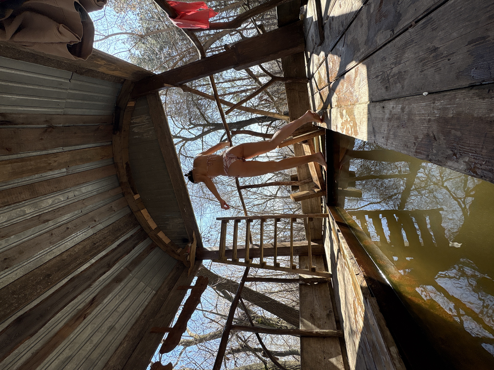 The image depicts the interior of a partially enclosed structure that appears to be built from wood and corrugated metal. The structure gives the impression of a makeshift or rustic sauna or bathhouse, perhaps located outdoors. A woman in a bikini is positioned on a set of wooden stairs or platform leading down to a pool of water. The water reflects the branches of trees above, suggesting the structure is in a wooded area.
While I don't have information to confirm a specific location in the city of Ulyanovsk, Russia, the design of the structure, the type of trees visible, and the practice of bathing in cold water suggest a location that could be in a similar climate or cultural context.