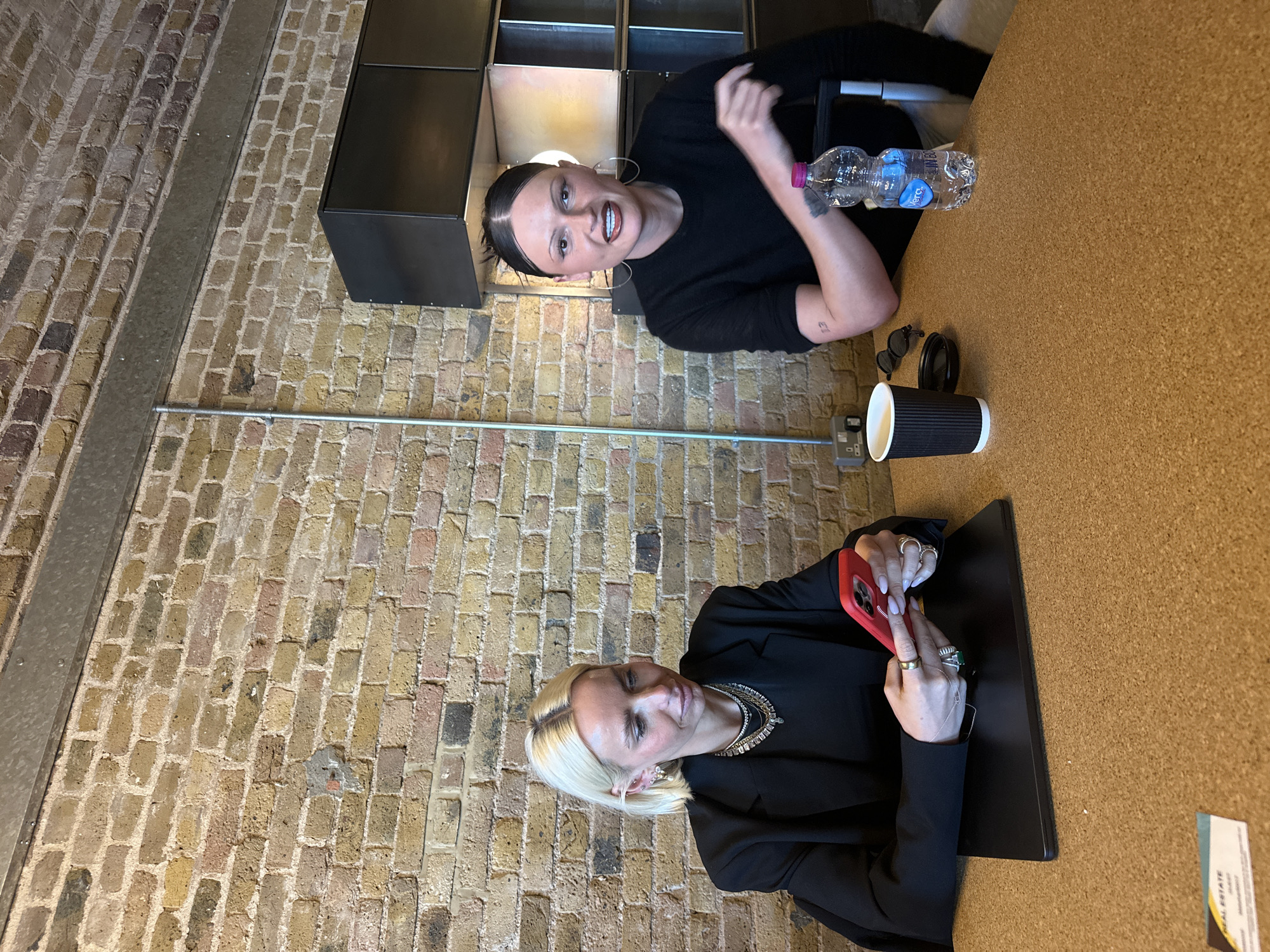 The image depicts two individuals seated at a cork-surfaced table within an indoor environment characterized by exposed multi-toned brick walls and galvanized steel structural elements, consistent with a converted commercial or industrial space in London, United Kingdom. The person on the left is a blonde woman wearing a black blazer and layered necklaces, engaged with a red smartphone held in both hands. A closed black laptop rests on the table in front of her. The person on the right has dark hair pulled back, wearing a black long-sleeved top and large hoop earrings. This individual is looking towards the camera with a slight smile and gesturing with their right hand. A small tattoo is visible on their left forearm. Objects on the table include a black ribbed paper coffee cup, black sunglasses, and a plastic water bottle with a pink cap and "Vera" label. A gray electrical outlet is visible on the brick wall to the left, and a business card showing "REAL ESTATE" is partially visible in the bottom-left foreground.