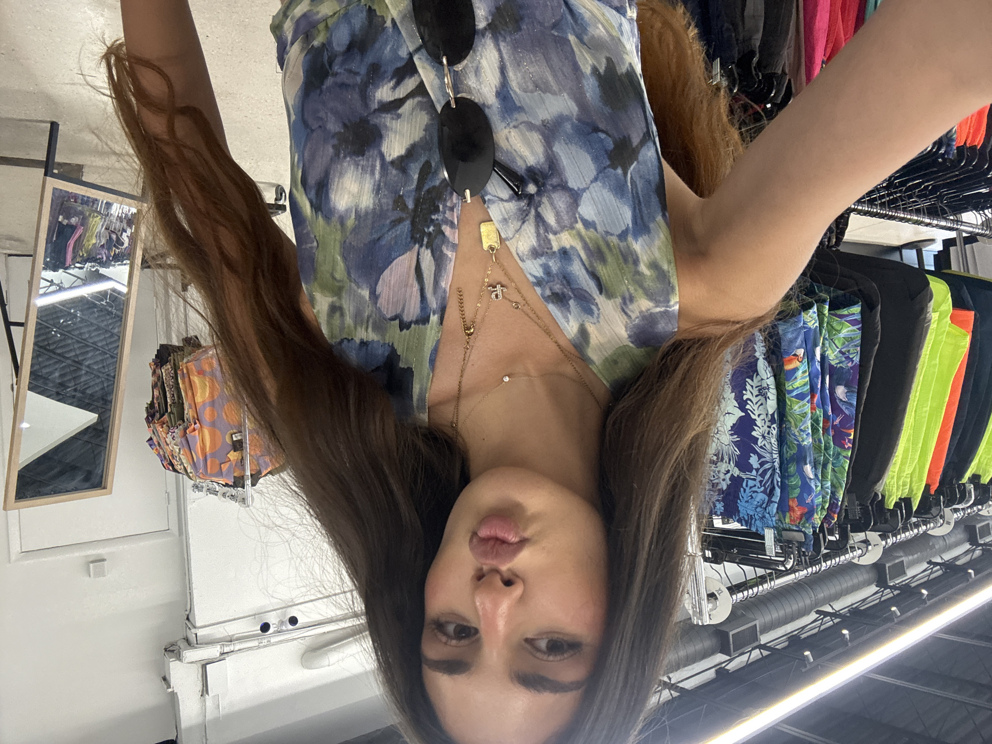 The image shows a woman with long brown hair in a store. She is wearing a blue floral print top and gold necklaces. Sunglasses are perched on her chest. The background shows clothing racks filled with colorful garments. The location is likely a clothing store in a modern setting with bright lighting and industrial-style ceiling fixtures. Based on the clothing styles, the location could be Miami, United States of America. There is a mirror reflecting the store's interior.