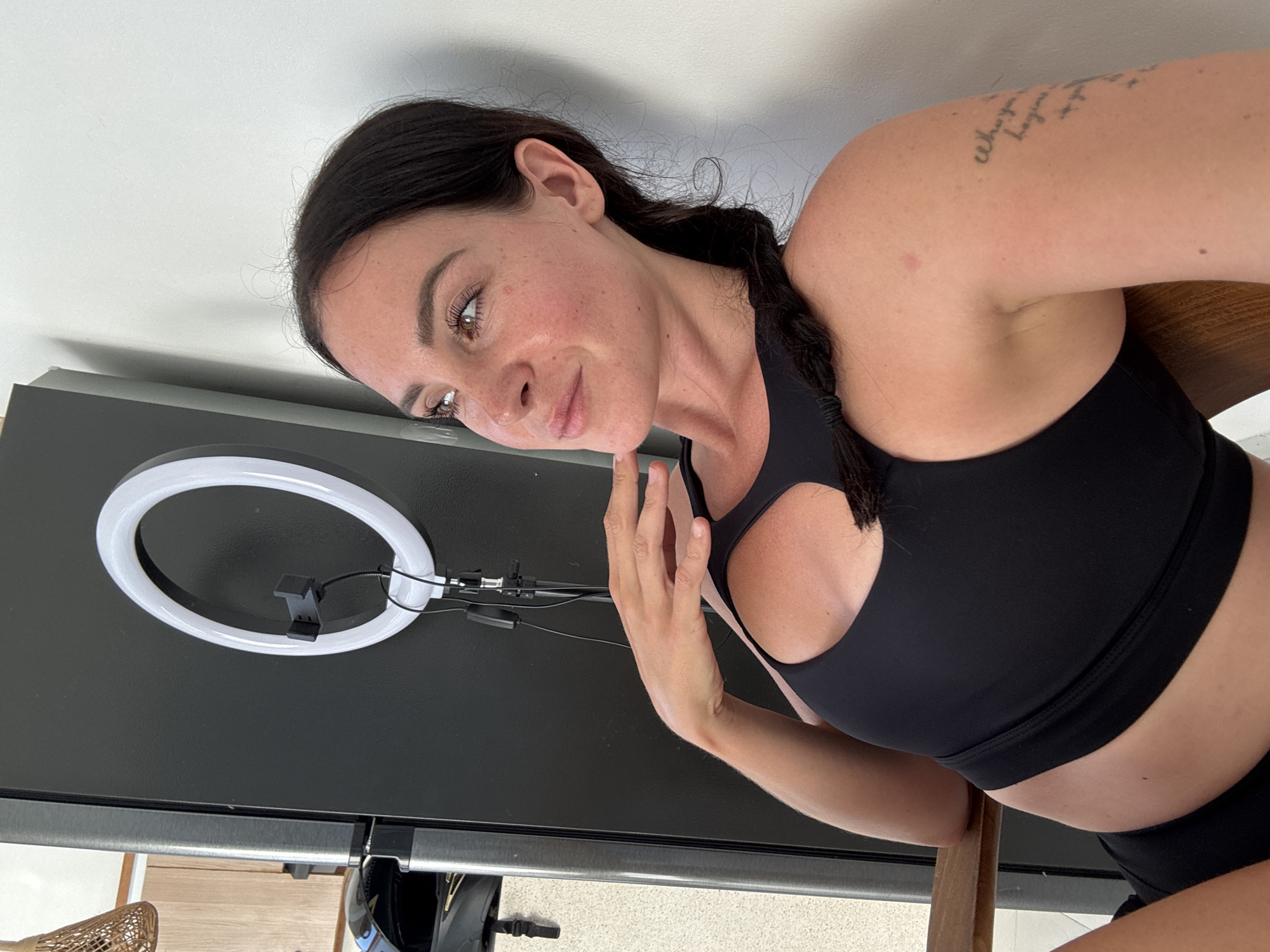 A woman with dark hair in a braid, wearing a black sports bra, is sitting in front of a black refrigerator with a ring light mounted on it. She is looking away from the camera, with her hand resting on her chin. The background is a plain white wall. The scene is indoors, likely in a home setting. The woman has freckles on her face and a tattoo on her arm.