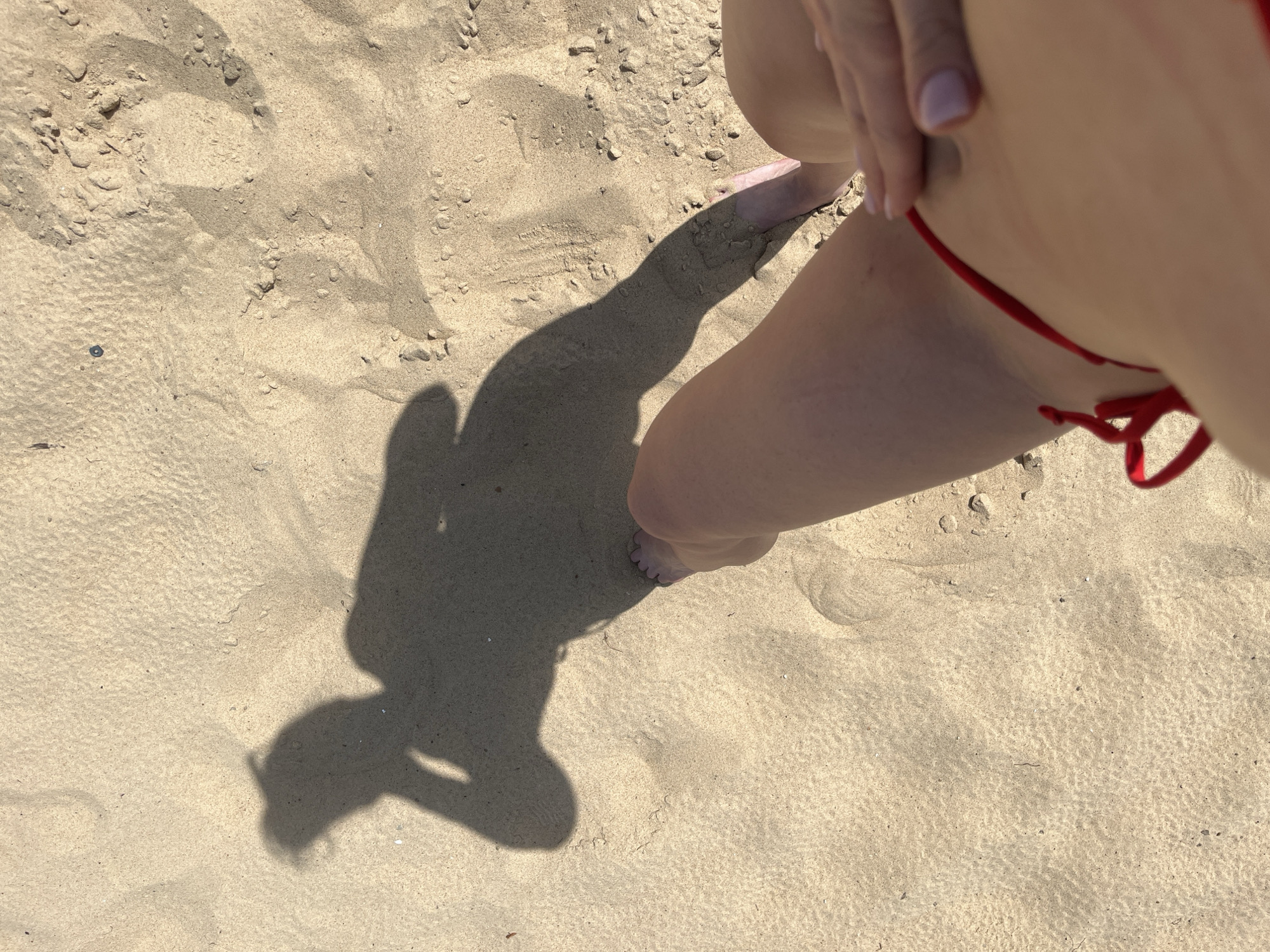 This is an overhead shot of a person's legs and shadow on a sandy beach. The person is wearing a red bikini bottom. The shadow shows a person with their arm raised to their head, suggesting they are posing for a picture. The sand is light brown and textured, with some small pebbles and debris scattered across it. The image does not contain any information that would allow identification of the location as Kazan, Russia.