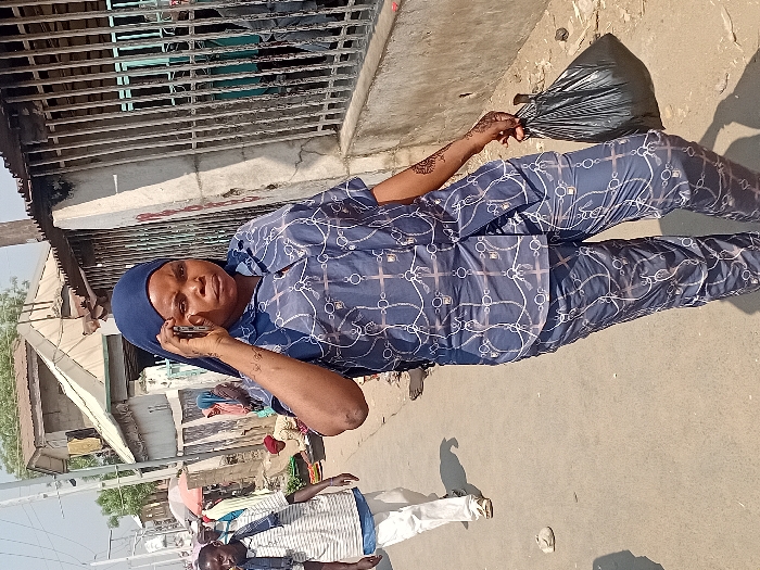 A woman in blue patterned pajamas and a headscarf stands on a dusty street, talking on a cell phone. She holds a black garbage bag. Two men walk nearby, one in a striped shirt and white pants. Behind them, a building with a metal grill overhangs part of the street. Other buildings and people are visible in the background. The scene is brightly lit.