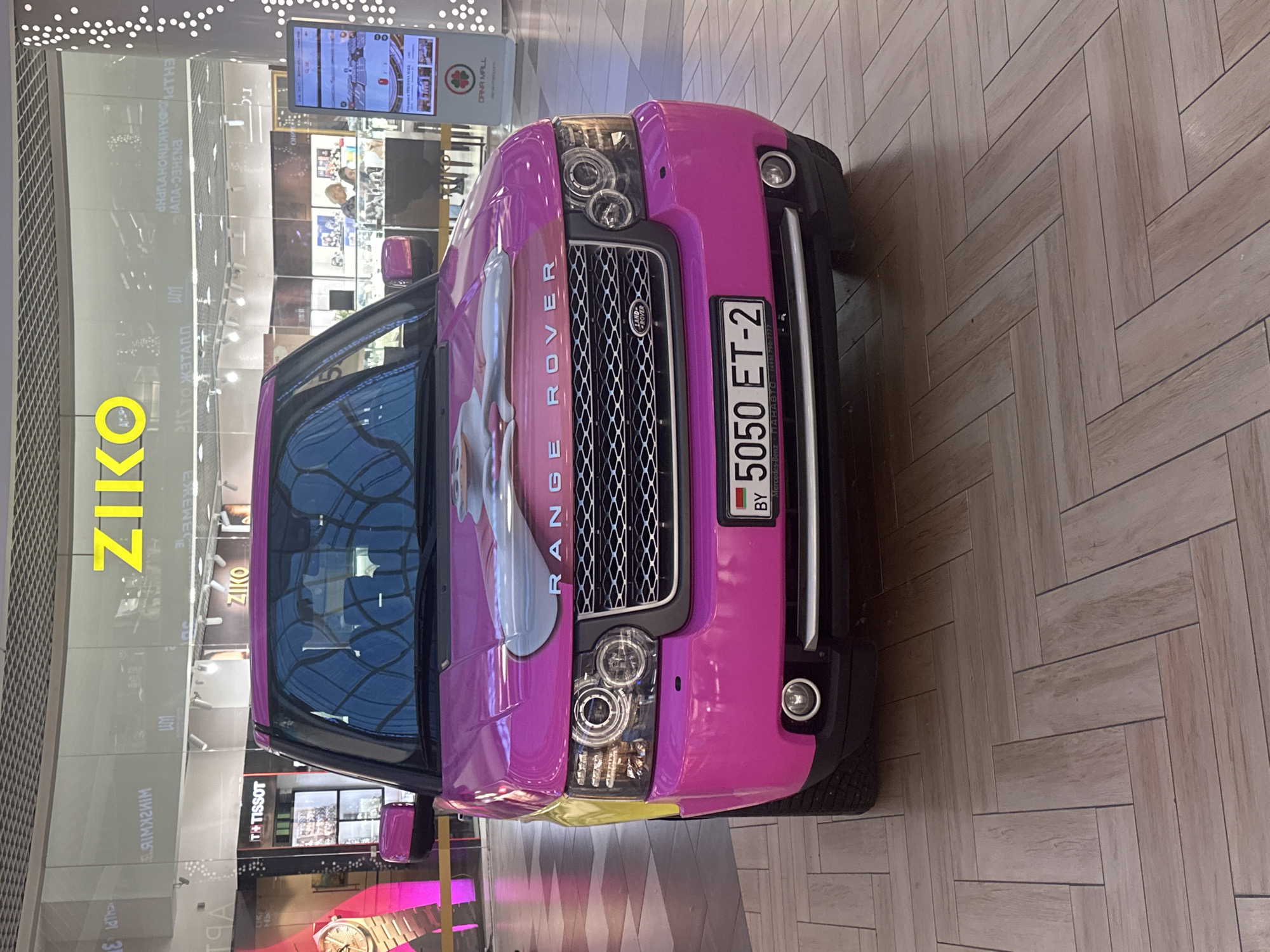 A bright pink Range Rover is parked indoors on a wooden floor in a shopping mall. The Range Rover has a decal on its hood depicting a cartoon character. The license plate on the car reads "5050 ET-2" and has "BY" indicating Belarus. In the background, there are shops with signs, including "ZIKO" and "TISSOT". Some people are visible in the distance, shopping or walking through the mall. The mall has a modern architectural design with large windows and a grid-like ceiling. The lighting suggests it is daytime. The overall scene depicts a commercial indoor space with a uniquely decorated vehicle as the focal point. The mall is located in Minsk, Belarus, as indicated by the license plate and the likely context of the shops displayed.