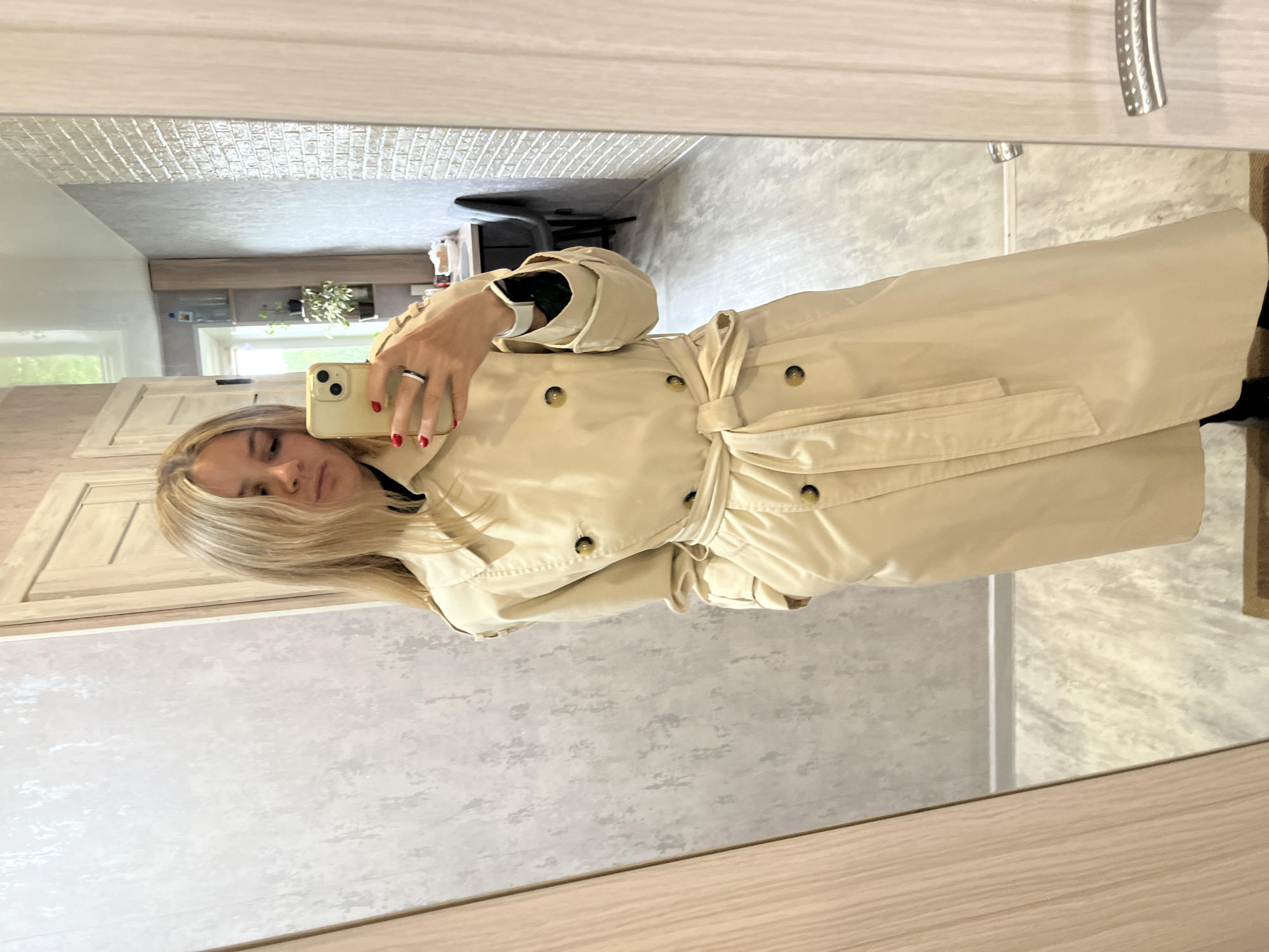 A woman with blonde hair takes a mirror selfie in a room. She is wearing a beige trench coat, a black top, and black pants. The room appears to be a hallway or entryway, with light-colored walls and wooden doors. In the background, a mirror reflects a shelf with plants and a table. The flooring is light wood. The setting is a residential interior. This image was taken on the street in Chaikovsky, Russia.