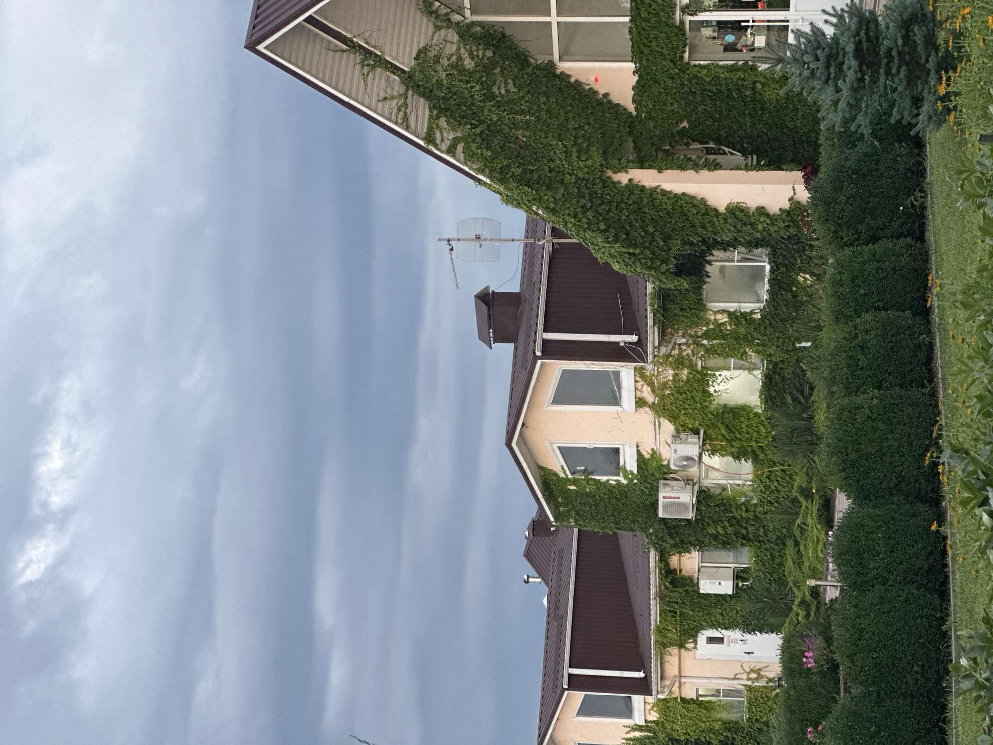The image shows a building with a brown roof, covered in ivy. The building has several windows and air conditioning units on the exterior walls. In the foreground, there are manicured green bushes and some yellow flowers. The sky is overcast with gray clouds. The location is identified as Yaroslavskaya, Russia.