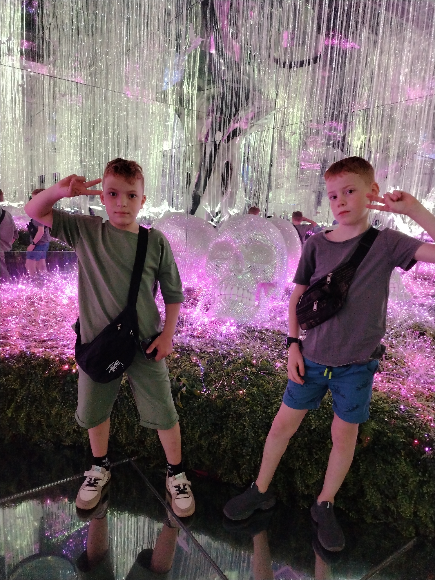 Two young, fair-skinned boys stand on a reflective surface, possibly glass, indoors. The boy on the left wears a light green t-shirt and matching shorts, black socks with white accents, and off-white sneakers. A black crossbody bag with a white logo is slung over his shoulder. He holds up a peace sign near his forehead. The boy on the right wears a gray t-shirt, blue shorts with a yellow drawstring, and black shoes. He also has a black crossbody bag and makes a peace sign.
Behind the boys is a decorative skull made of shimmering material, glowing with purple light. The background consists of hanging silver strands that resemble rain or beaded curtains. The ground is covered in lush green vegetation interspersed with twinkling purple lights.
The location is the Kazan Family Center in Kazan, Russia.