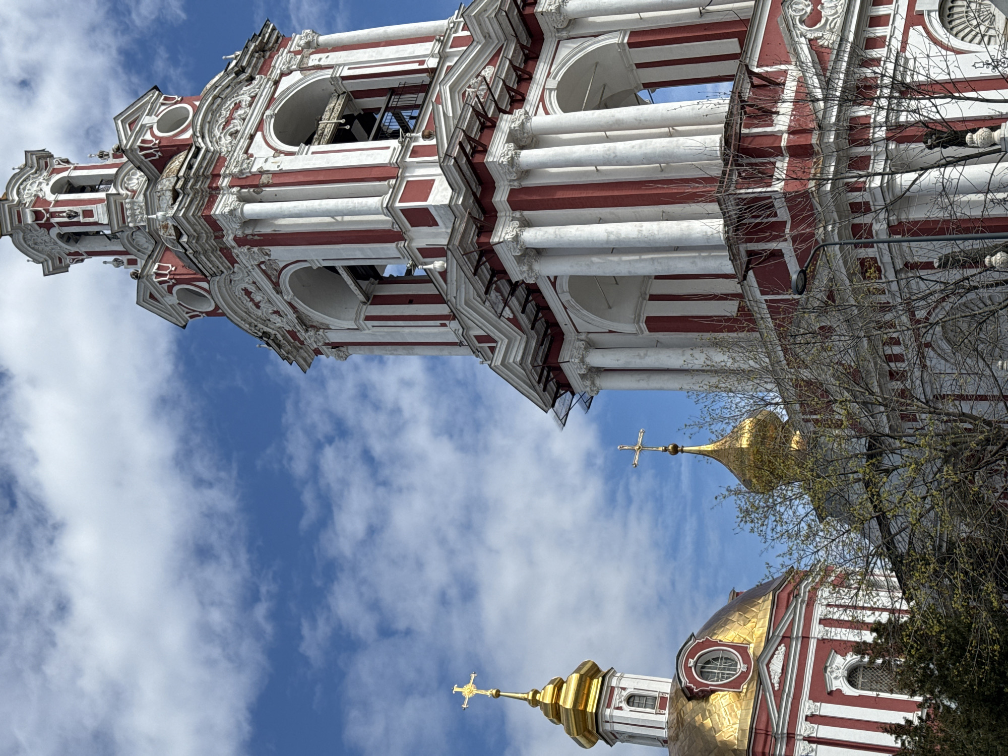 The image shows a low-angle view of the Novodevichy Convent in Moscow, Russia, including a red and white bell tower and golden-domed cathedrals against a partly cloudy blue sky. The architecture features ornate details, arches, and columns. Tree branches with budding leaves are also visible.