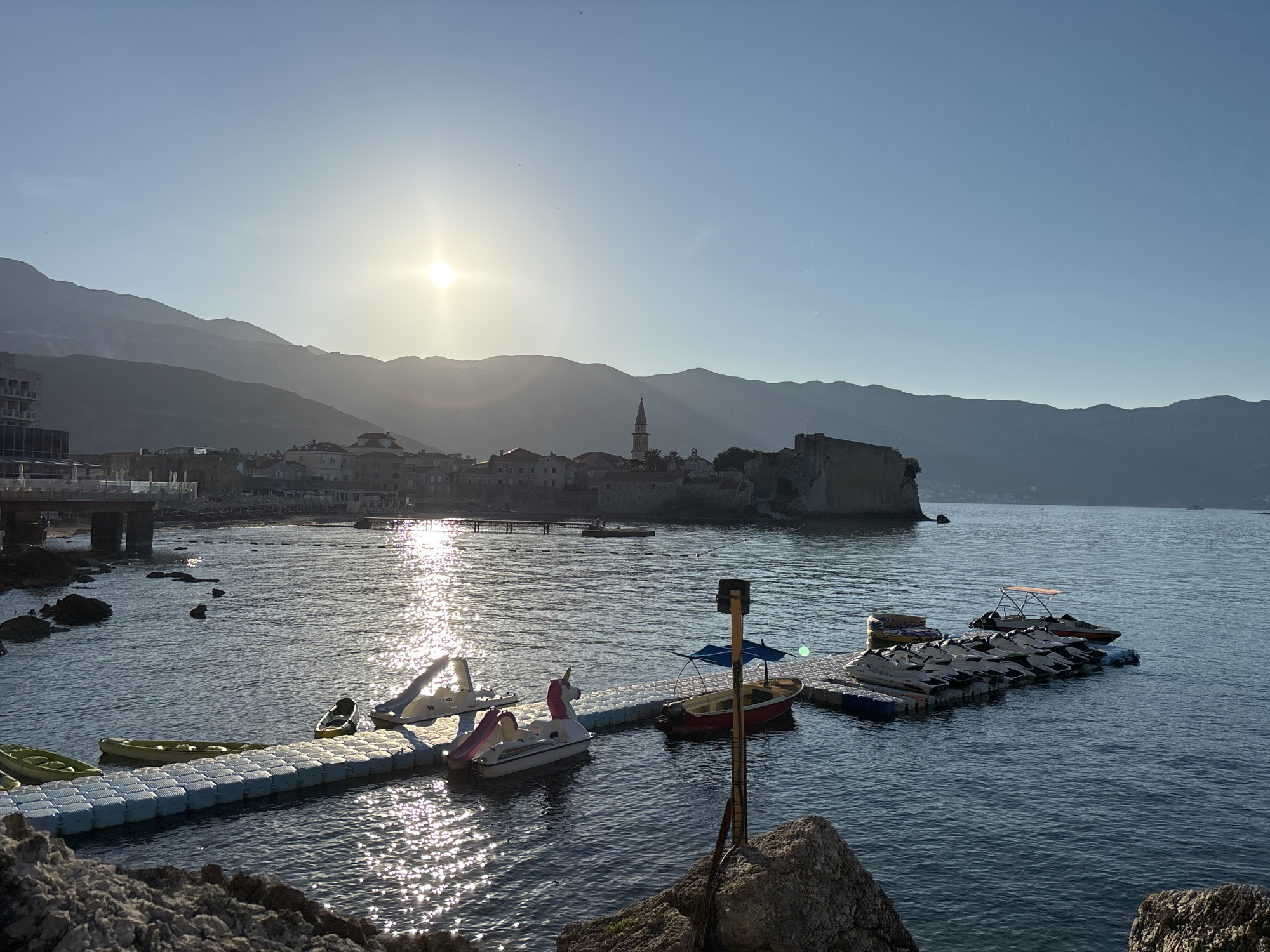 The image depicts a coastal scene at sunrise or sunset, with the sun low on the horizon, casting a warm glow and creating shimmering reflections on the water. In the foreground, a rocky shoreline leads to a modular floating dock with several watercraft. These include kayaks, a pedal boat with a unicorn design, a small red boat with a canopy, and a row of jet skis. The water appears calm, with gentle ripples.
In the mid-ground, a town with red-tiled roofs is visible along the coast, dominated by a prominent church steeple and a historic fortress or citadel perched on a rocky outcrop. Behind the town, a range of mountains forms a majestic backdrop, their peaks softened by the atmospheric light. The sky is a clear, pale blue, with a few distant, faint birds visible. The overall impression is one of serene natural beauty and a hint of human activity in a picturesque coastal setting. The location is identified as Unknown, Unknown.