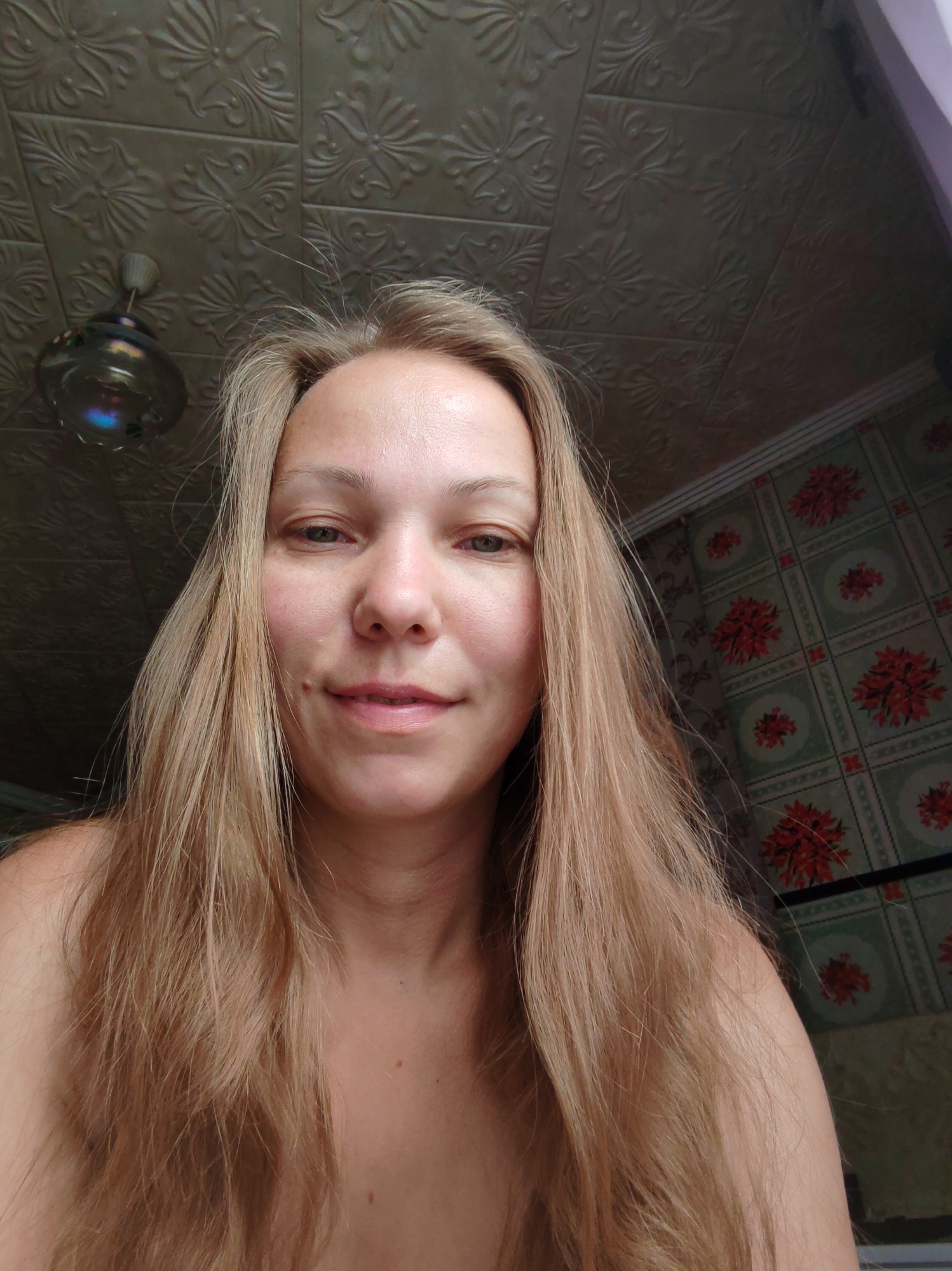 A young woman with long blonde hair is pictured from the chest up, looking directly at the camera. She has fair skin and is smiling slightly. The background shows a patterned ceiling with a decorative fixture and wallpaper with a floral design. The scene appears to be indoors.