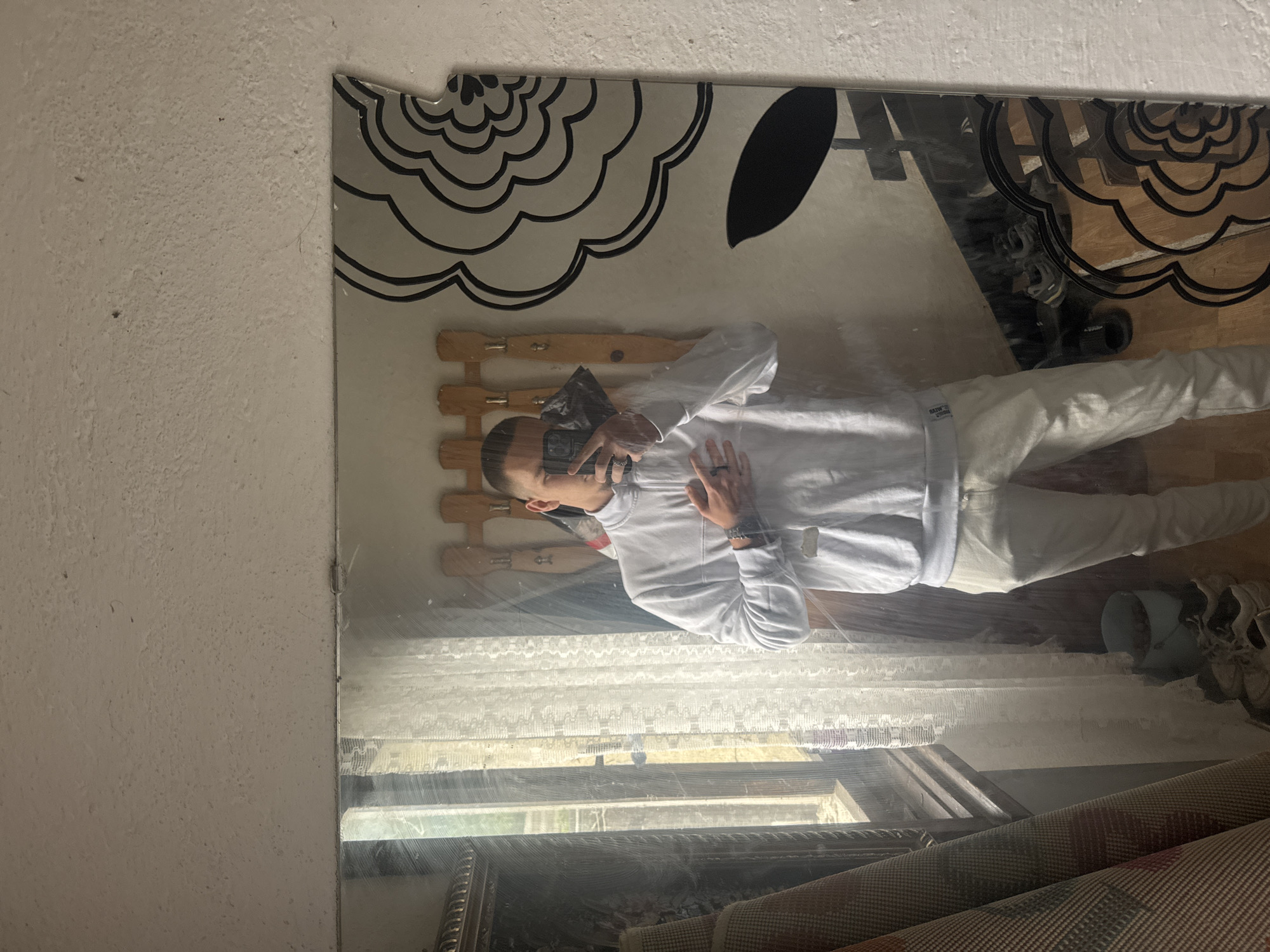 A man is taking a selfie in a mirror. He is wearing a white sweatshirt and light-colored pants. The mirror reflects a cluttered room with a coat rack, a window, and rolled-up rugs. The mirror itself has a decorative black and white pattern on its top edge. The location is Vetren Dol, Bulgaria.