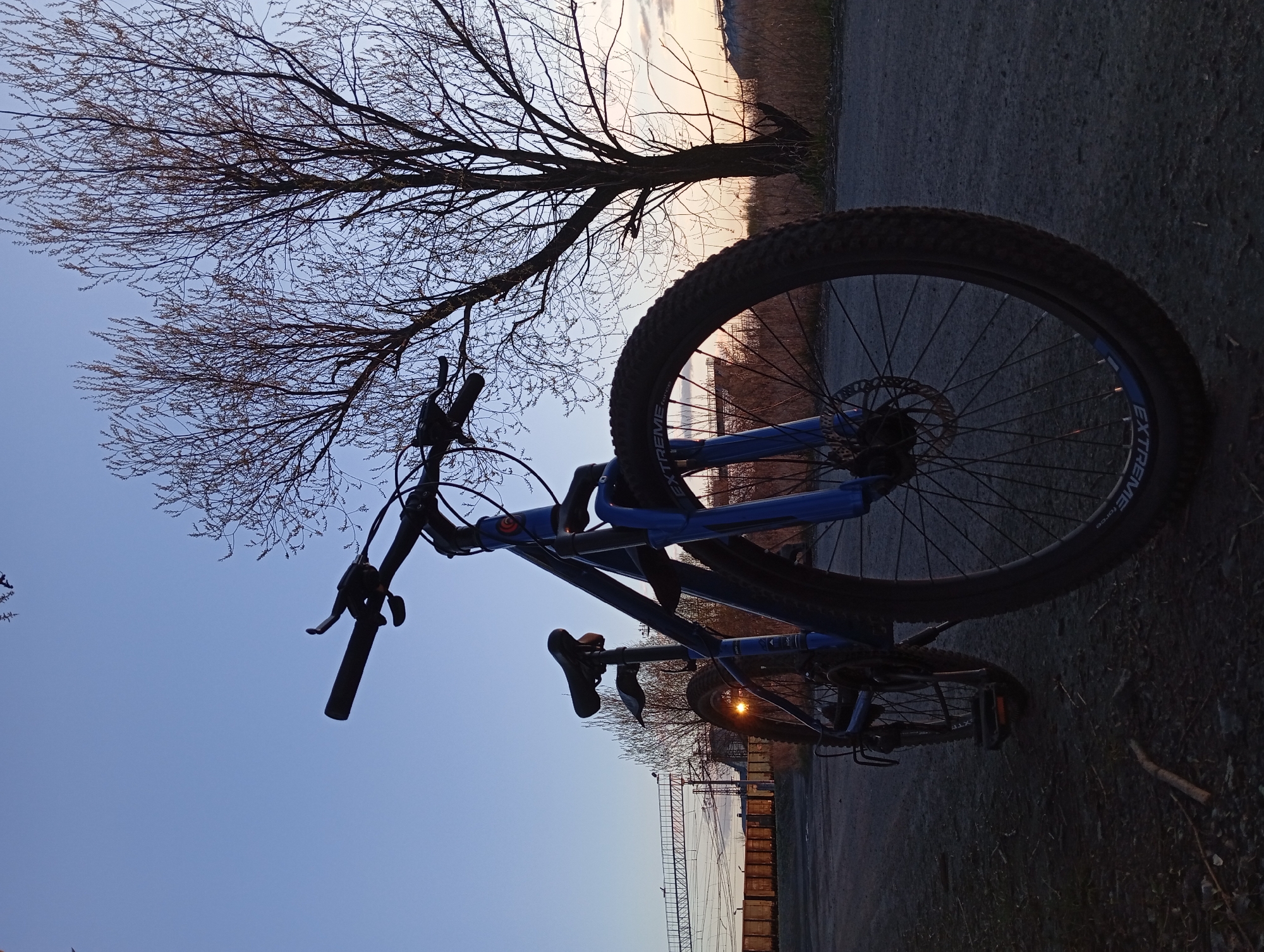 A bicycle is silhouetted against the early morning sky and landscape. The bicycle is blue and black. It is parked on an asphalt path next to a bare tree, with a view of a fence and the railway tracks of the Yurgamysh train station. The sky is pale blue, tinged with orange and yellow from the sunrise. The scene is likely in or near Yurgamysh, Russia, near the train station.