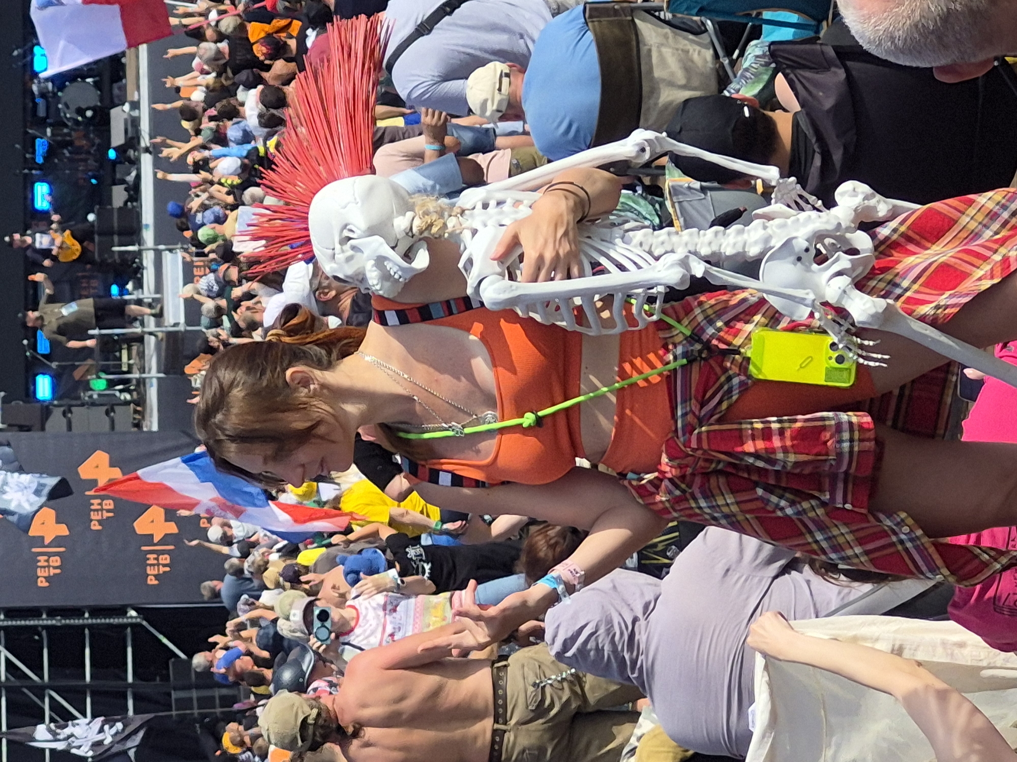 The image depicts a crowded outdoor music festival in Lagunovo, Russia. In the foreground, a woman with brown hair, wearing an orange crop top and a red and yellow plaid shirt tied around her waist, holds a plastic skeleton prop with red hair spikes. She is looking to the right. Behind her, a large crowd of people are visible, many with their hands raised. In the background, a stage with musical equipment and performers can be seen. Flags and banners with logos are displayed, including "PEH TB". The overall atmosphere suggests a lively event with a large audience.