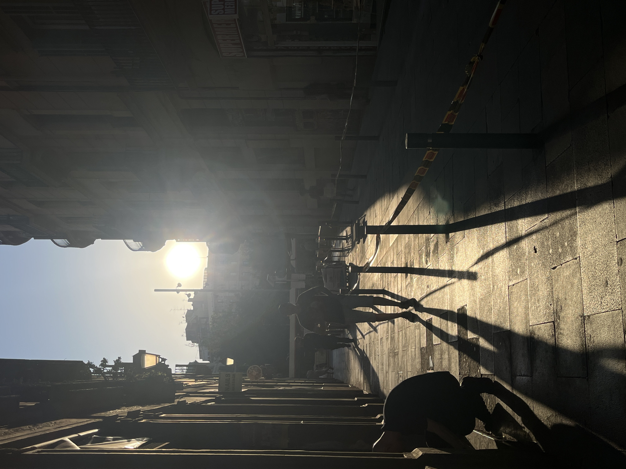The image shows a street scene with pedestrians walking in a city, location unknown. The photo is taken from a low angle, looking up towards the sun. The sun is high in the sky, casting long shadows of the people and objects on the ground. The buildings on either side of the street are tall and block out some of the light. The street is made of pavement. A metal barrier is visible on the side of the road.