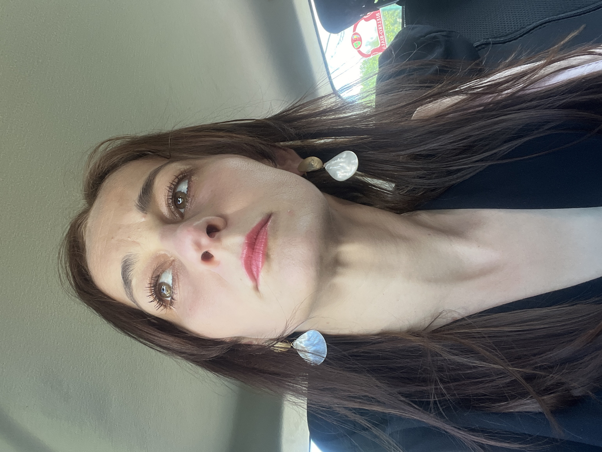A close-up, medium shot captures a woman from the chest up, looking upward and to the left. She has long, dark brown hair, styled with bangs, and is wearing a black jacket. Her face is made up with lipstick, and she has large, dangling earrings with white, translucent shells. The background appears to be the interior of a vehicle, with a grey headliner and a window visible on the right side. A sticker with Polish text is adhered to the window. The lighting suggests an outdoor daytime setting, with sunlight illuminating the woman's face.