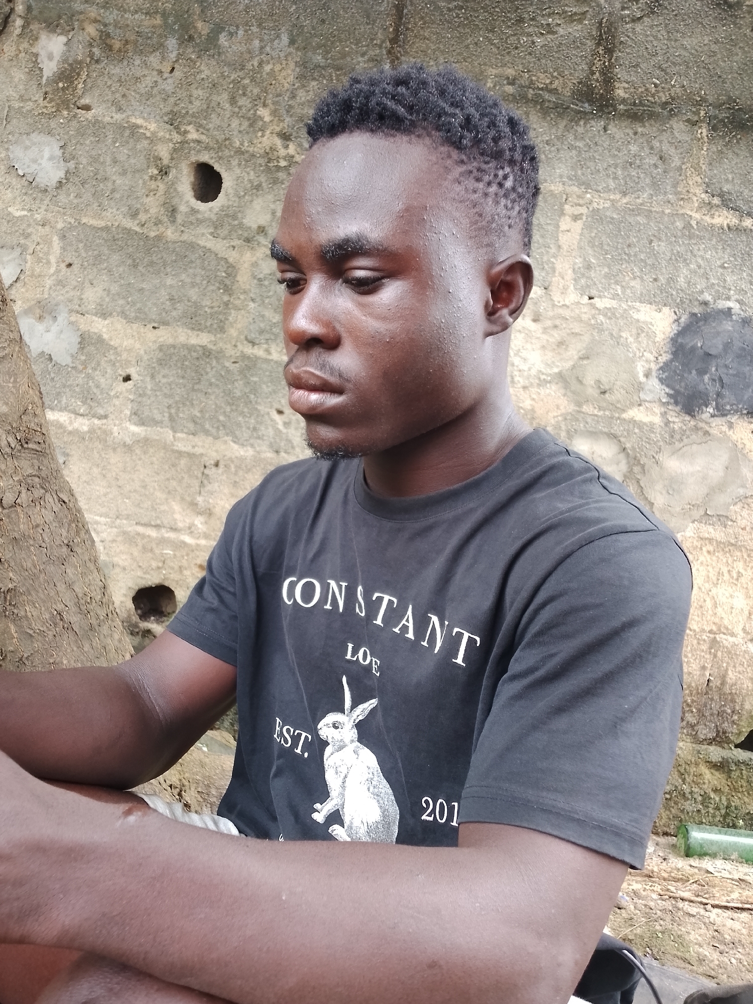 The image shows a young Black man sitting outdoors in front of a rough, concrete wall. He is wearing a black t-shirt with white text and a graphic of a rabbit. The man is looking down and to his left. His arms are resting on his lap. To his left, a tree trunk is visible. The ground around him is dirt and some debris. The wall behind him has several holes and discolored patches. A green glass bottle is visible on the ground to the right. The scene appears to be in a somewhat neglected urban area, consistent with parts of Ado, Nigeria.