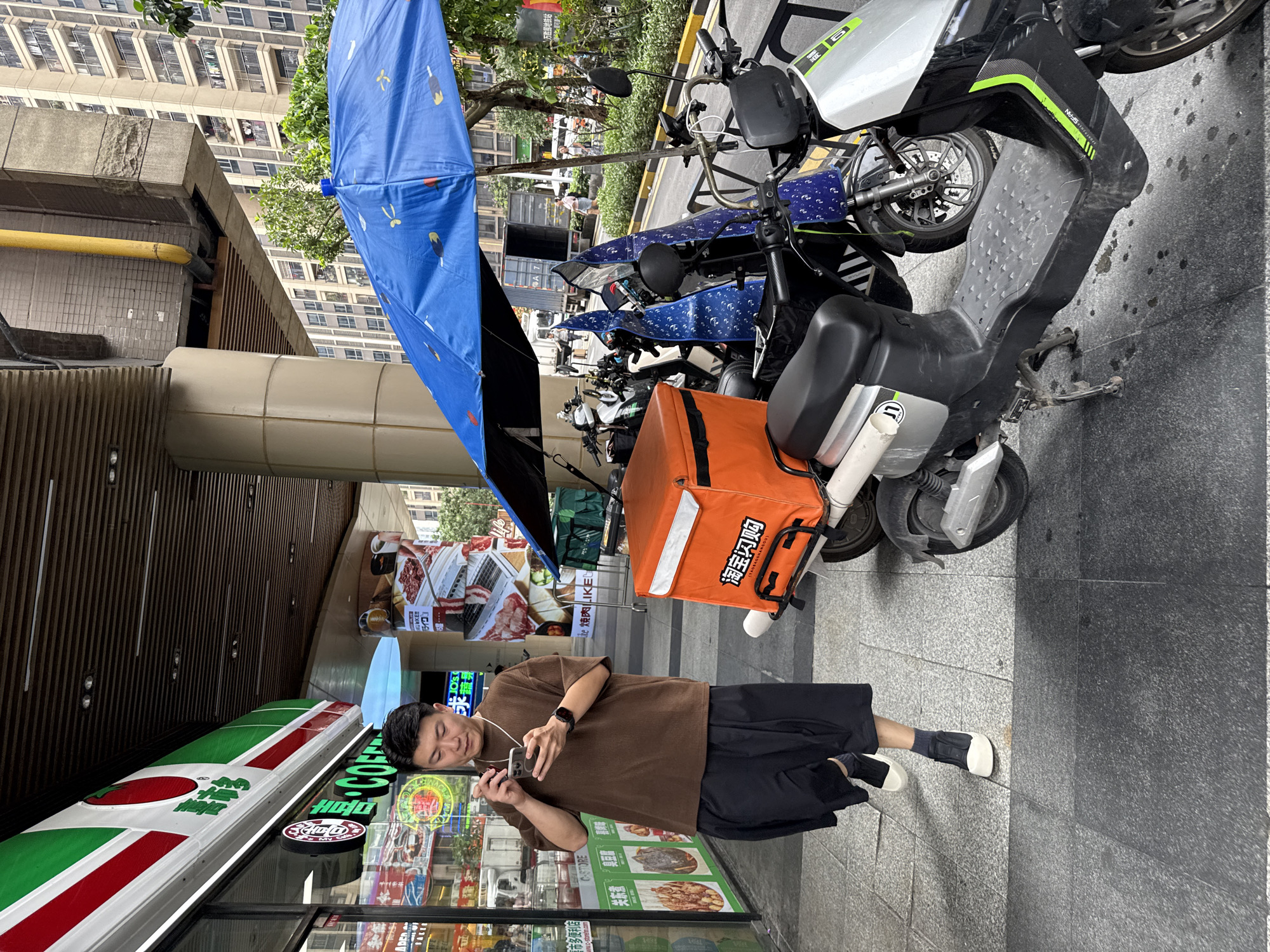 A man in a brown t-shirt and black shorts walks on a tiled sidewalk next to a row of parked electric scooters. He is looking down at his phone, which he holds in his right hand. A large blue umbrella with a patterned design is open above some of the scooters. One scooter in the foreground has an orange insulated delivery box attached to the back. Behind the scooters, a building with multiple stories and balconies is visible, along with trees and vegetation. The scene appears to be a street in Haizhu District, China, with shops visible on the left.