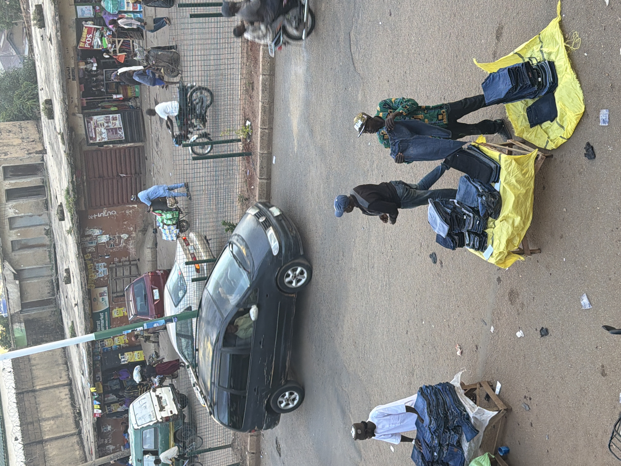 This is a vertical, eye-level, outdoor shot of a street market scene in Bukuru, Nigeria. The scene depicts a busy marketplace with several individuals engaged in commerce.
In the foreground, two men are observed arranging stacks of blue jeans on yellow fabric spread on wooden pallets. One man, wearing a patterned long-sleeved shirt and a cap, is standing and appears to be sorting through a pile of jeans. Another man in a white shirt and dark cap is also present, with a large stack of jeans in front of him.
Further into the scene, a black minivan is parked on the street. Inside the minivan, a person can be seen through the tinted windows. Beyond the minivan, a few other vehicles, including a red car and a white van, are visible, along with what appears to be a market stall with various goods displayed.
Several people are seen walking or standing near the market stalls. One person in a light blue shirt is pushing a cart loaded with items. In the background, there are buildings and fences, suggesting an urban environment.
The image is taken during daylight, likely in the morning or afternoon, with no indication of specific weather conditions. The overall atmosphere seems to be one of everyday commerce and activity in a bustling Nigerian street market. The quality of the image is fair, with some areas appearing slightly out of focus. No visible text is clearly decipherable.