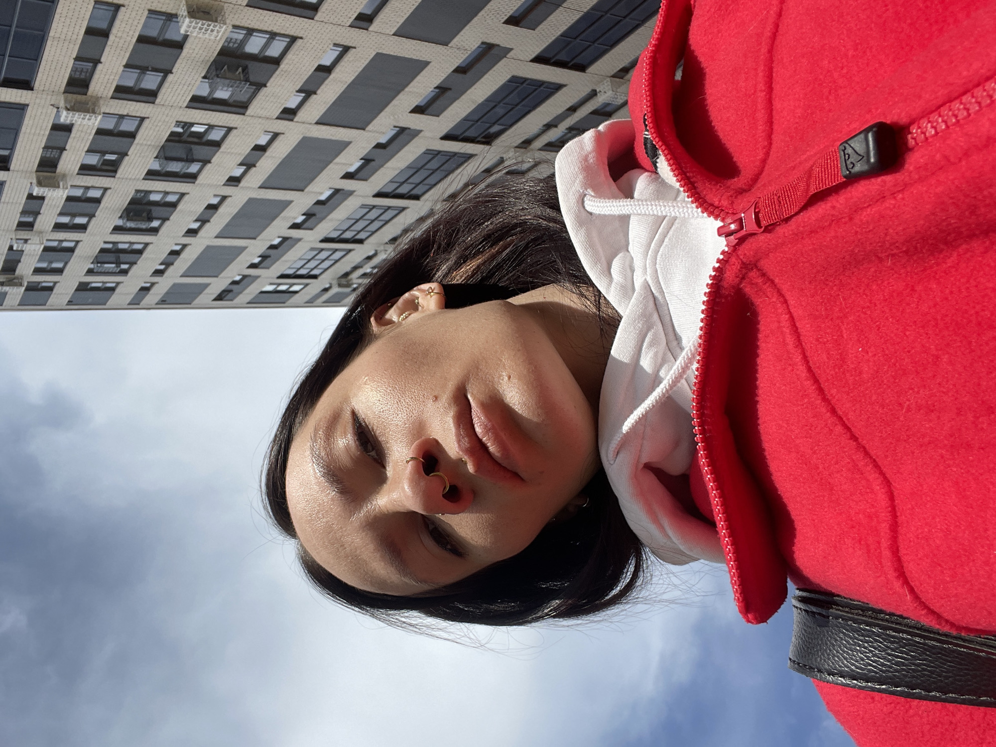 The image depicts a low-angle, close-up view of a female individual against an urban background in Izhevsk, Russia. The subject, positioned centrally, presents a downward gaze towards the camera. She has dark hair and visible facial piercings, including a septum ring and a left nostril ring, along with multiple gold-colored earrings in her right ear. Her skin shows a subtle sheen. She is dressed in a white hooded sweatshirt layered under a bright red zippered jacket, a black strap from a bag or backpack visible on her left shoulder. The upper left of the frame features a partially cloudy blue sky. To the right and upper right, a modern, multi-story building is visible, characterized by a light-colored facade with alternating rectangular windows and dark panels. Several external air conditioning units are mounted on the building's exterior.