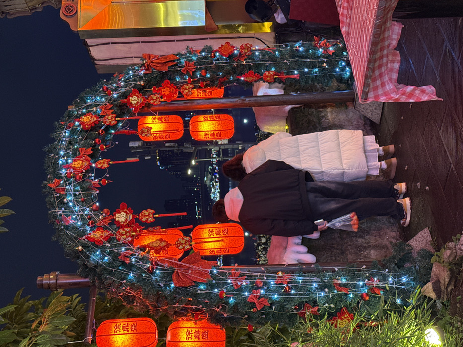 The image depicts a nighttime scene in Yuzhong District, China, focused on an illuminated festive archway. The archway is constructed from evergreen material, wrapped with blue string lights, and heavily adorned with numerous red paper lanterns, red bows, and intricate red Chinese knot-style ornaments featuring the character "福" (fú, meaning good fortune). The lanterns emit a warm orange glow and display Chinese characters vertically.
Two individuals stand beneath the archway, facing away from the viewer towards a city skyline visible in the distant background. The individual on the left is wearing a dark jacket with a light-colored hood, dark pants, and multi-colored athletic shoes. This individual holds a clear plastic bag containing several small orange fruits in their left hand. The individual on the right wears a long, white quilted coat, a white dress or skirt beneath it, and light brown flat shoes. Both individuals are observing the illuminated city structures and a dark body of water visible beyond the archway.
To the left of the archway, green foliage is visible, with a small, circular light source near the base. To the right, a table draped with a red and white checkered tablecloth is partially visible in the foreground. Irregular white shapes are situated behind the two individuals, possibly ground-level decorations or structures. The overall scene is brightly lit by the archway decorations and ambient city lights, contrasting with the dark night sky.